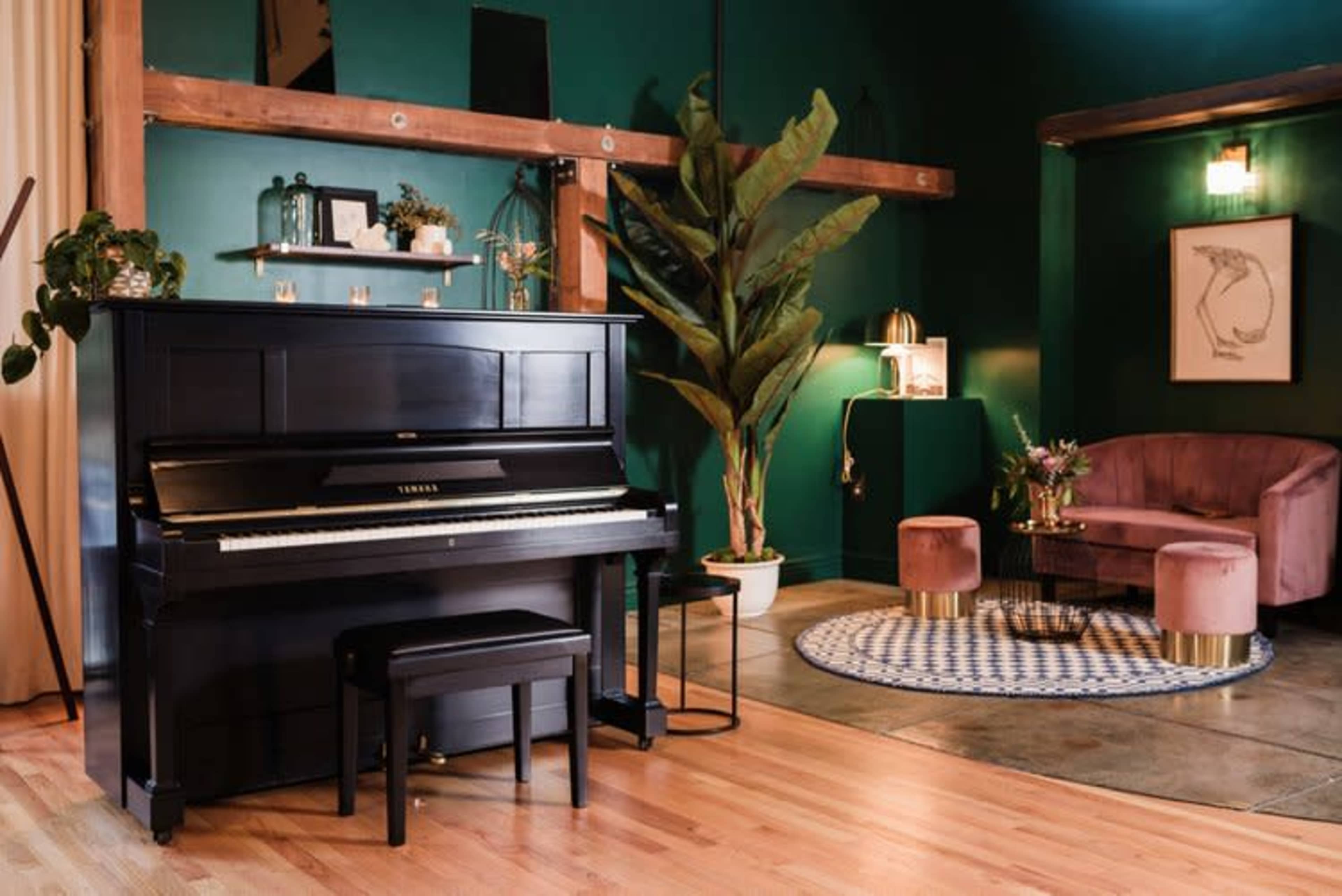 A black upright piano sits beside a plush purple seating area in a room with dark green walls and wooden beams.
