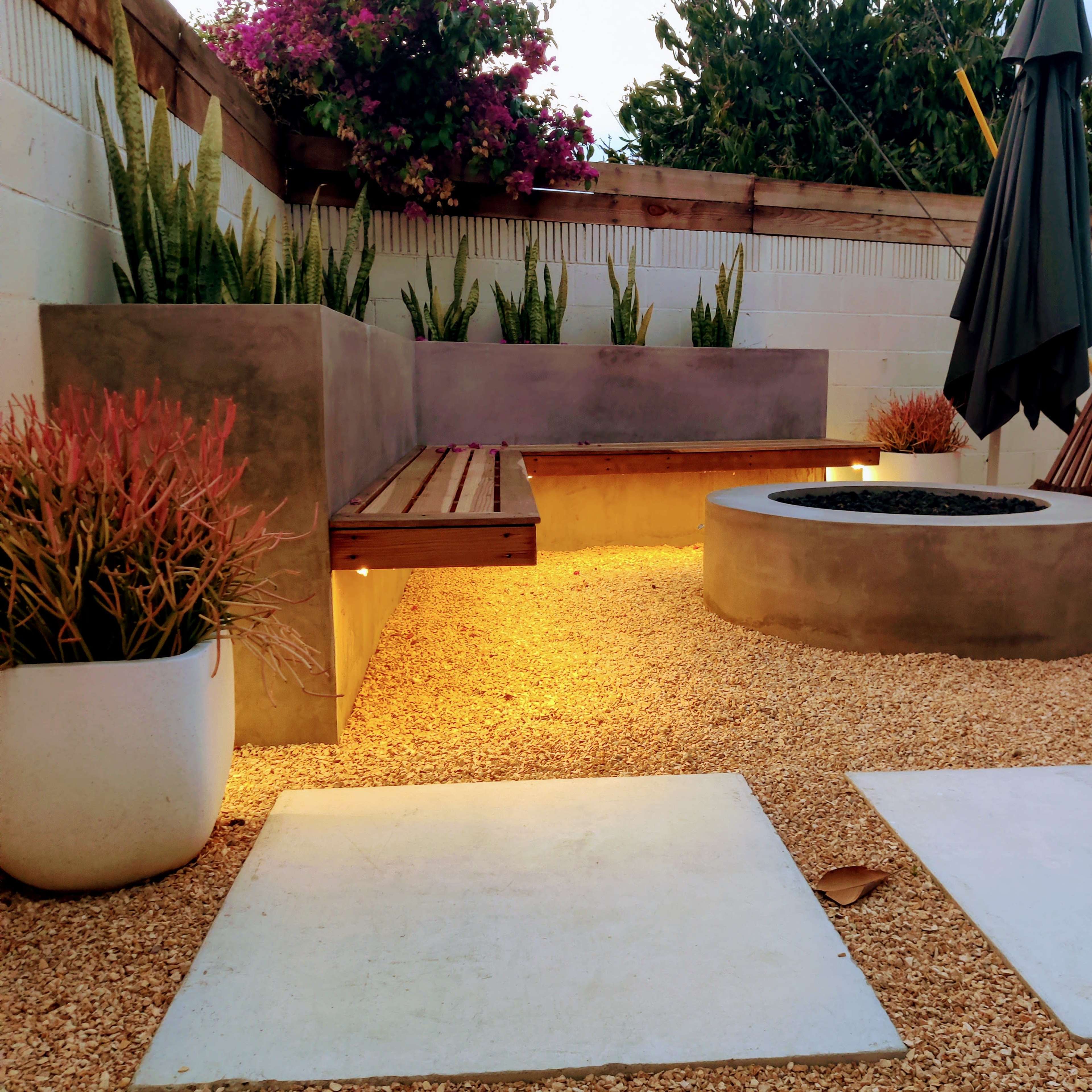 A landscaped outdoor space features a concrete seating area, a circular fire pit, and modern planters, illuminated by soft lighting.