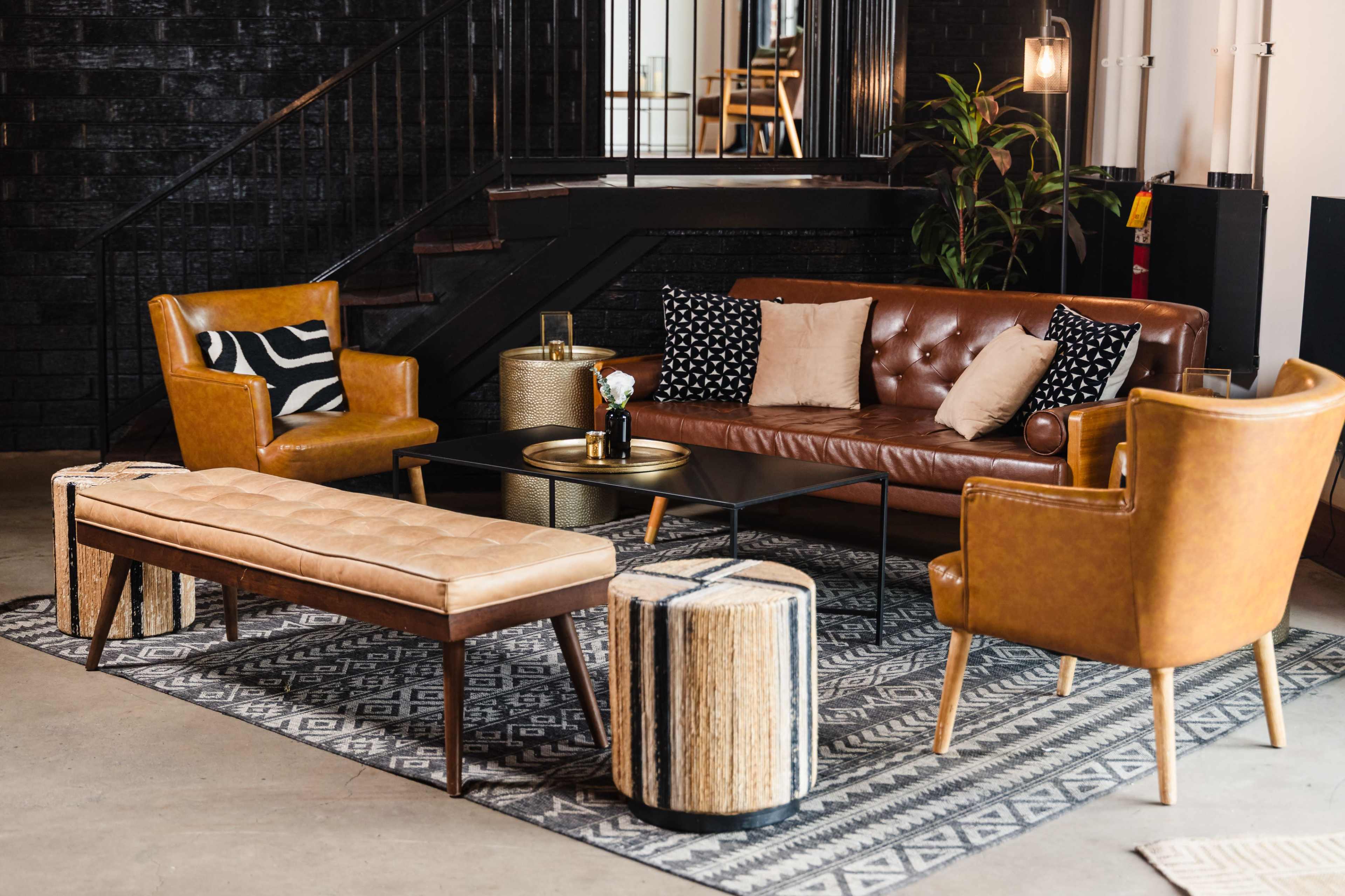 The image shows a modern living room setup featuring a brown leather sofa, two mustard-yellow armchairs, a round stool, and a rectangular bench, arranged on a patterned rug in front of a staircase.
