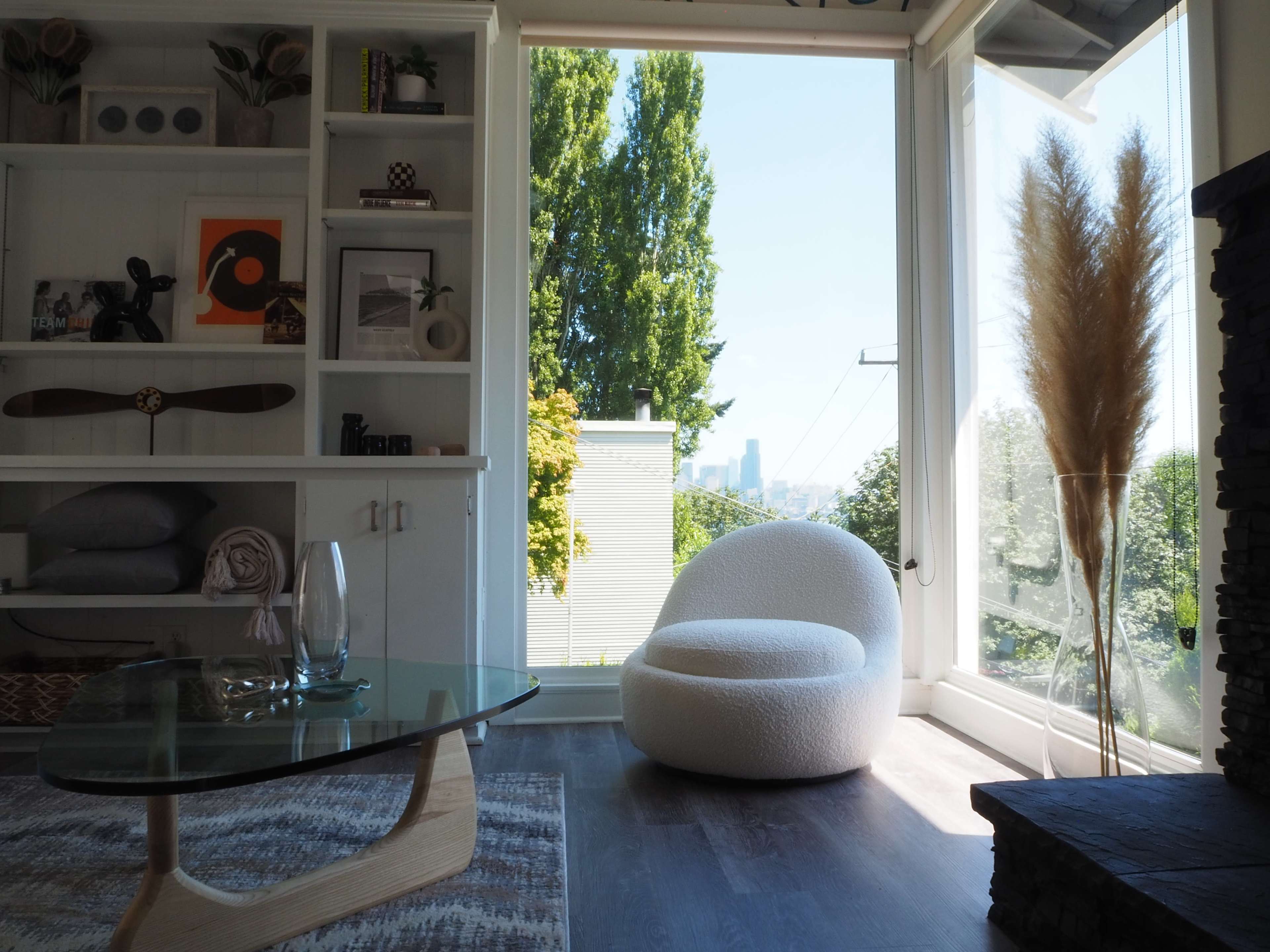 A modern living room with a round white chair, a glass coffee table, and a large window that offers a view of greenery and a city skyline.