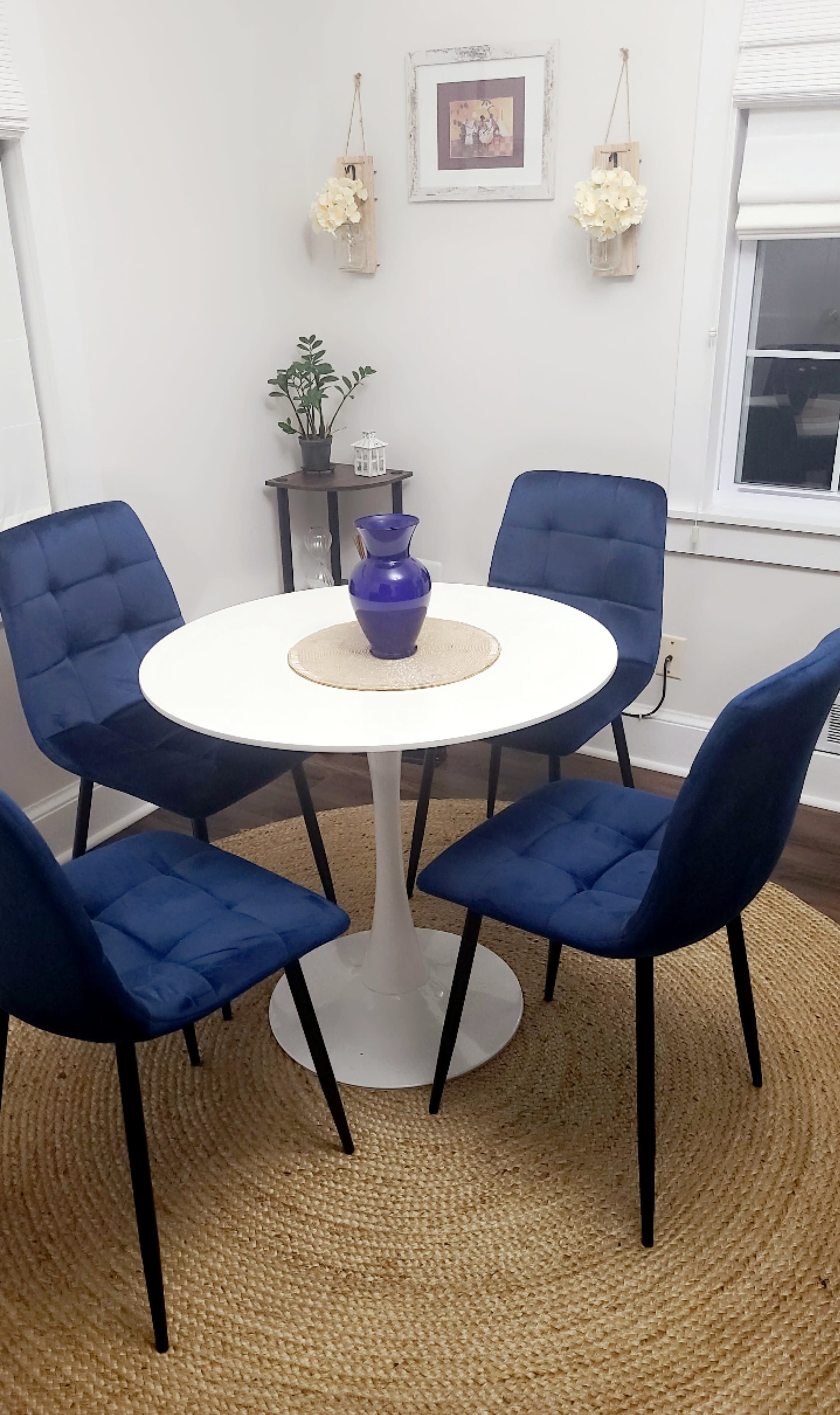 A round white table with four blue upholstered chairs is set on a woven rug in a well-lit dining area, complemented by a vase on the table and decorative items on the wall.