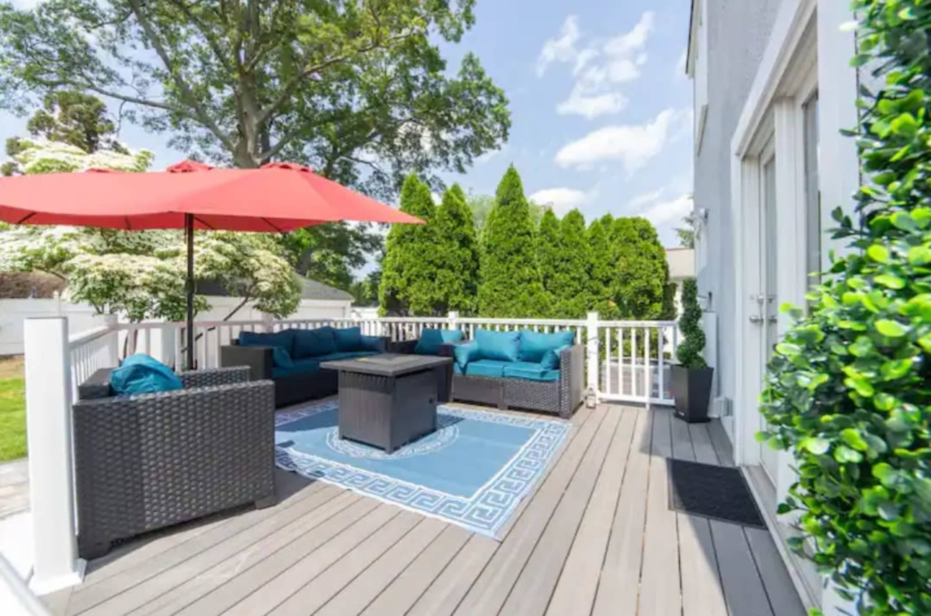 A modern outdoor deck features a seating area with dark wicker furniture, bright blue cushions, a central coffee table, and a red umbrella for shade.
