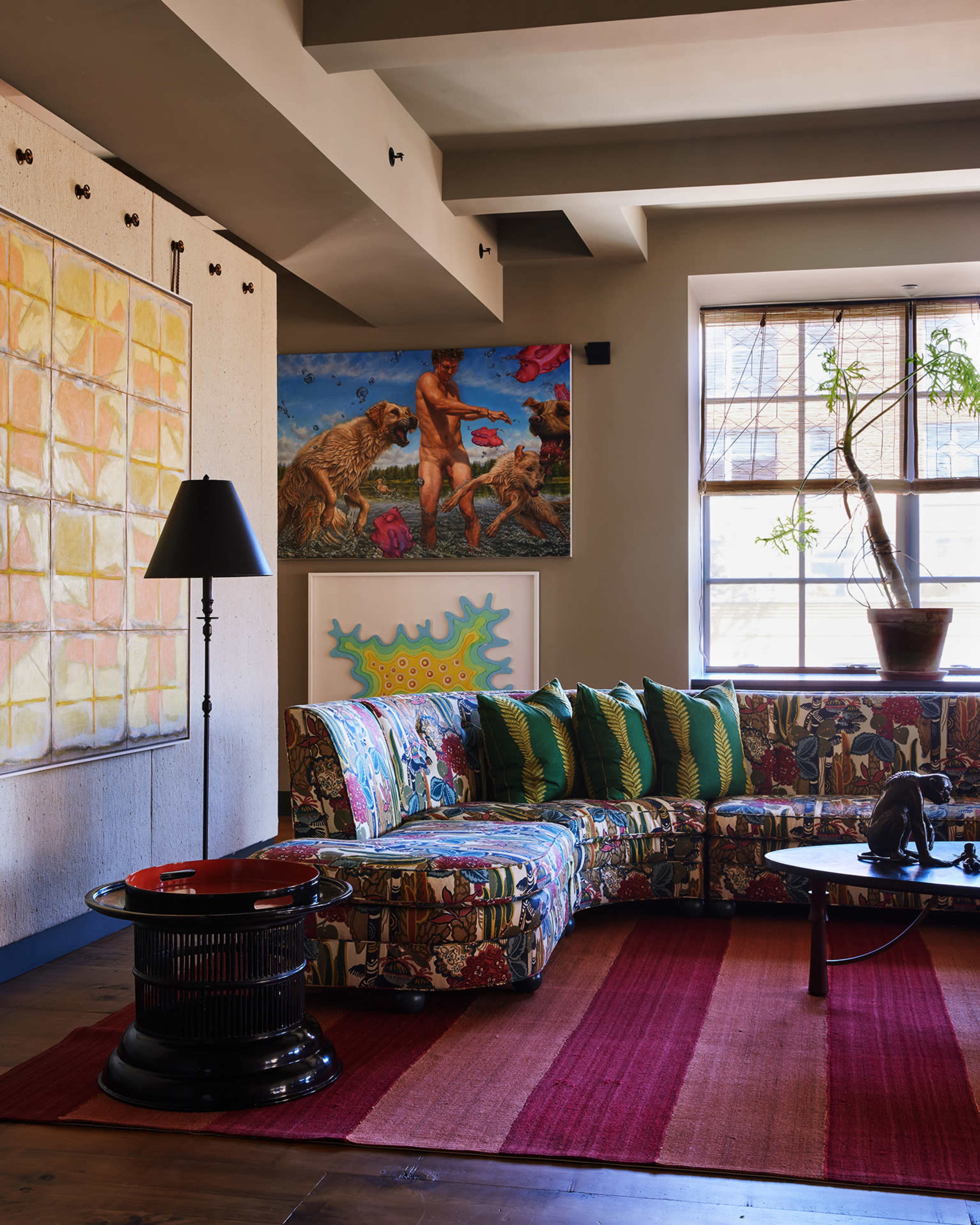 The scene features a colorful, patterned sofa in a living room with various artworks on the walls, including a large painting and a potted plant by the window.