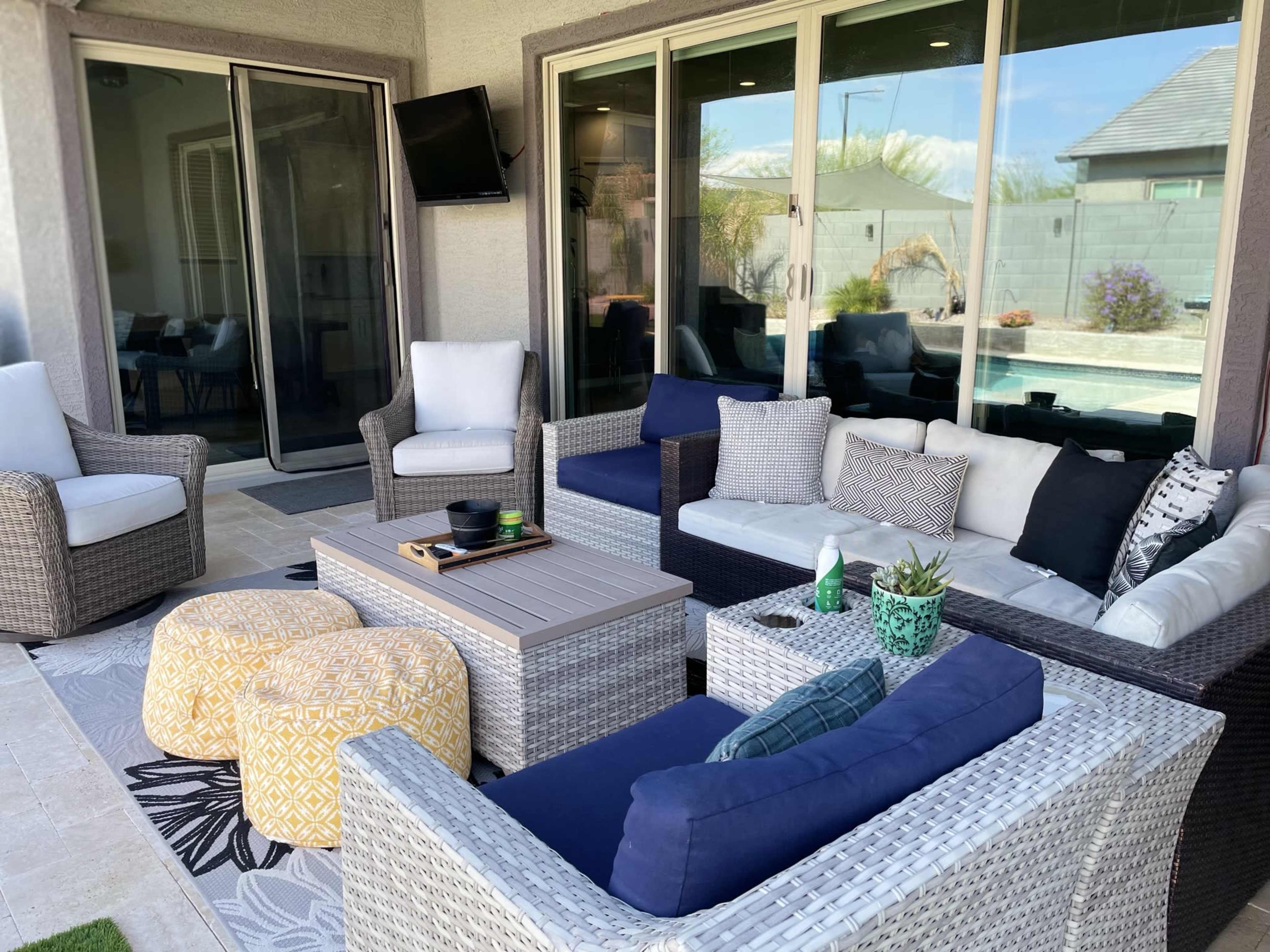 A modern outdoor seating area features wicker furniture, colorful cushions, and a central table, surrounded by large windows that overlook a pool.