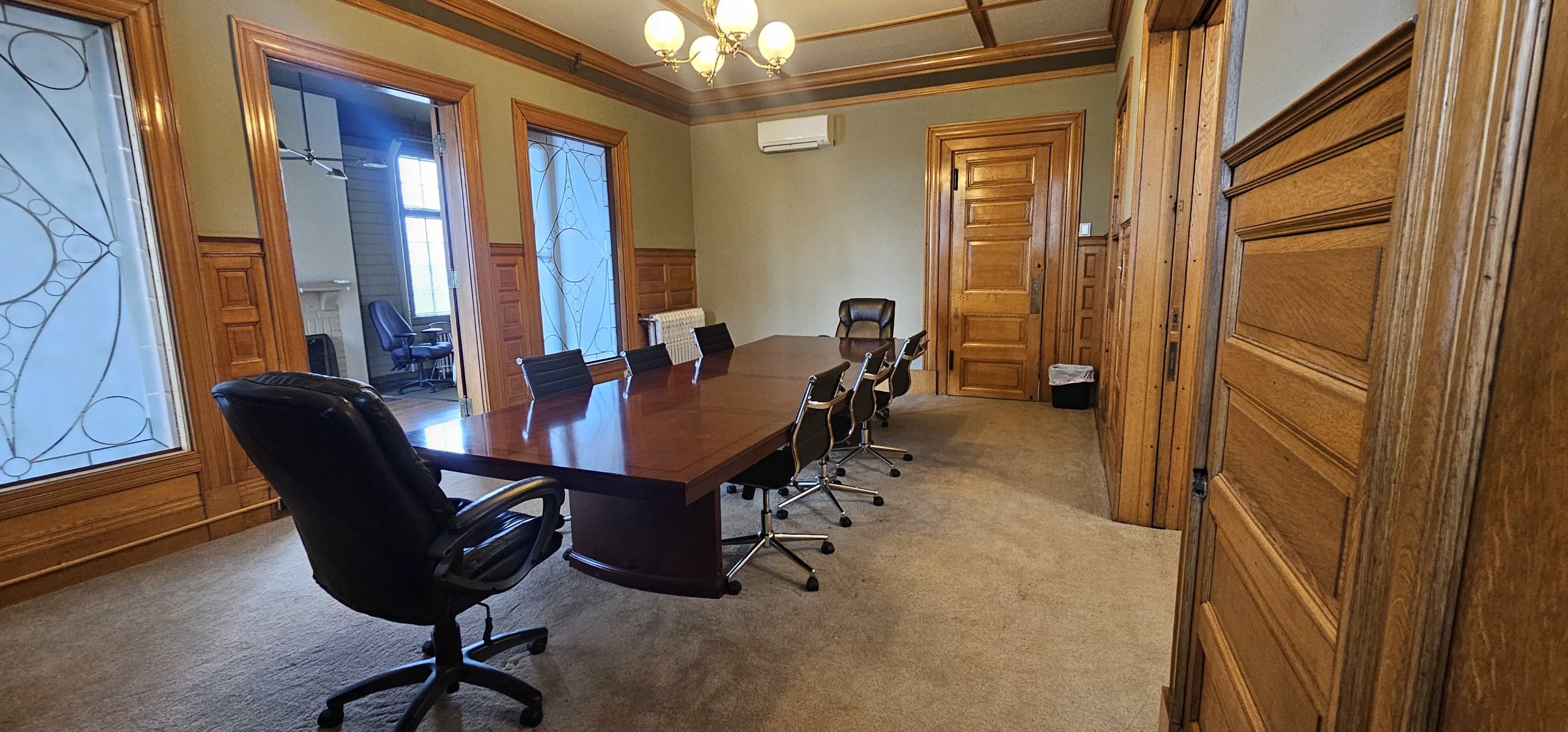 A conference room featuring a large wooden table surrounded by black chairs, with wooden paneled walls and large windows.