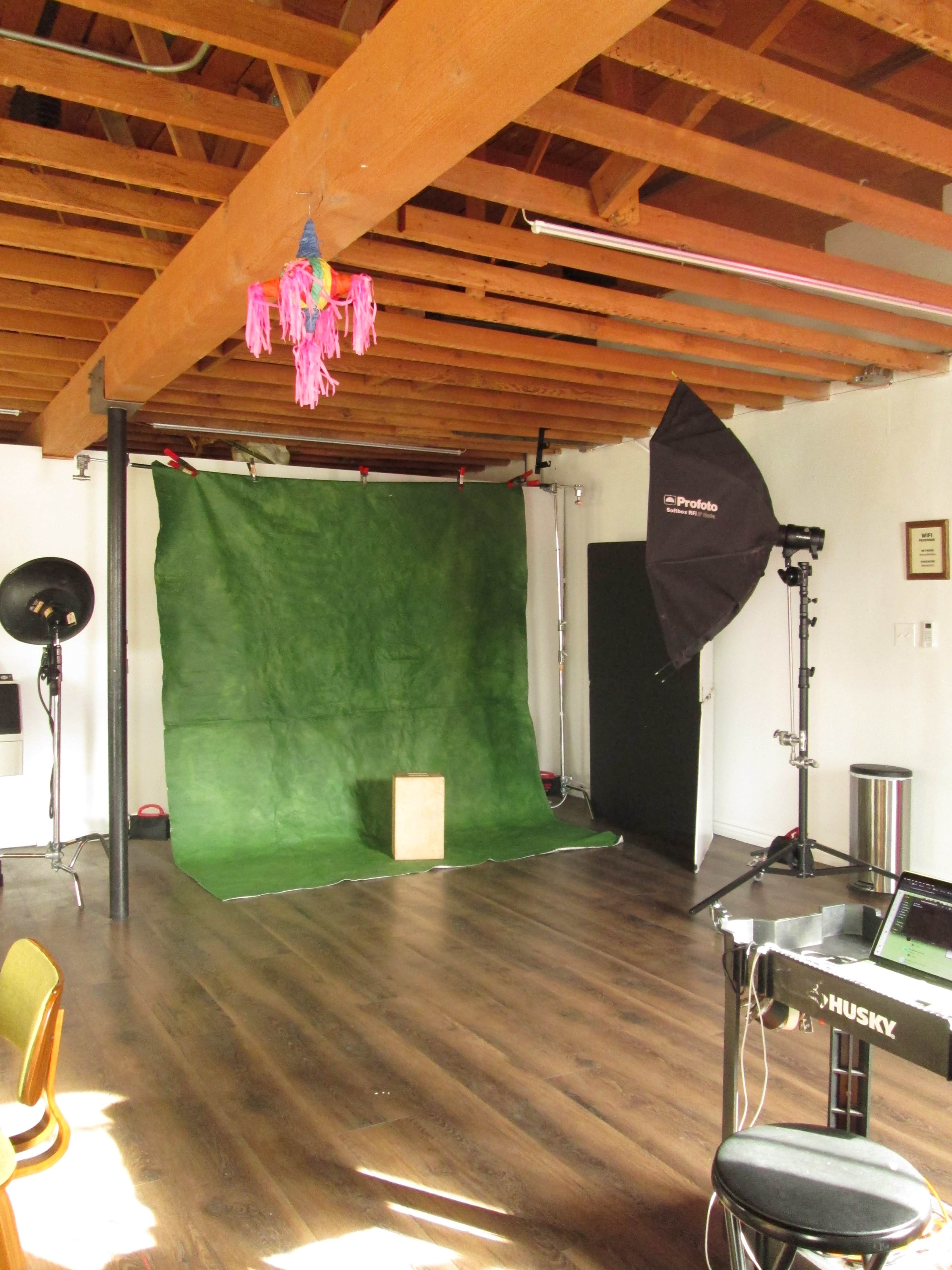 The image shows a photography studio with a green backdrop, a light stand, a photography accessory on the floor, and wooden beams overhead.