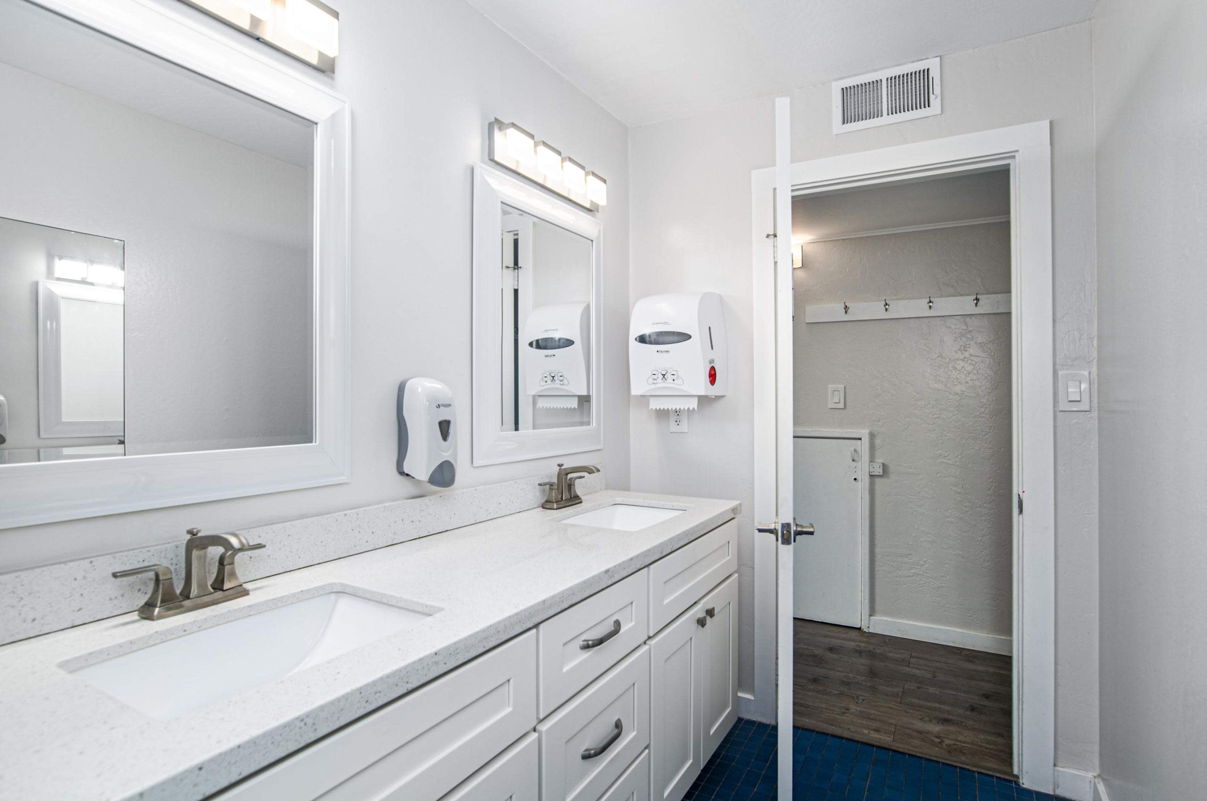 A bright, modern bathroom features two sinks, wall-mounted mirrors, and a separated area visible through an open door.