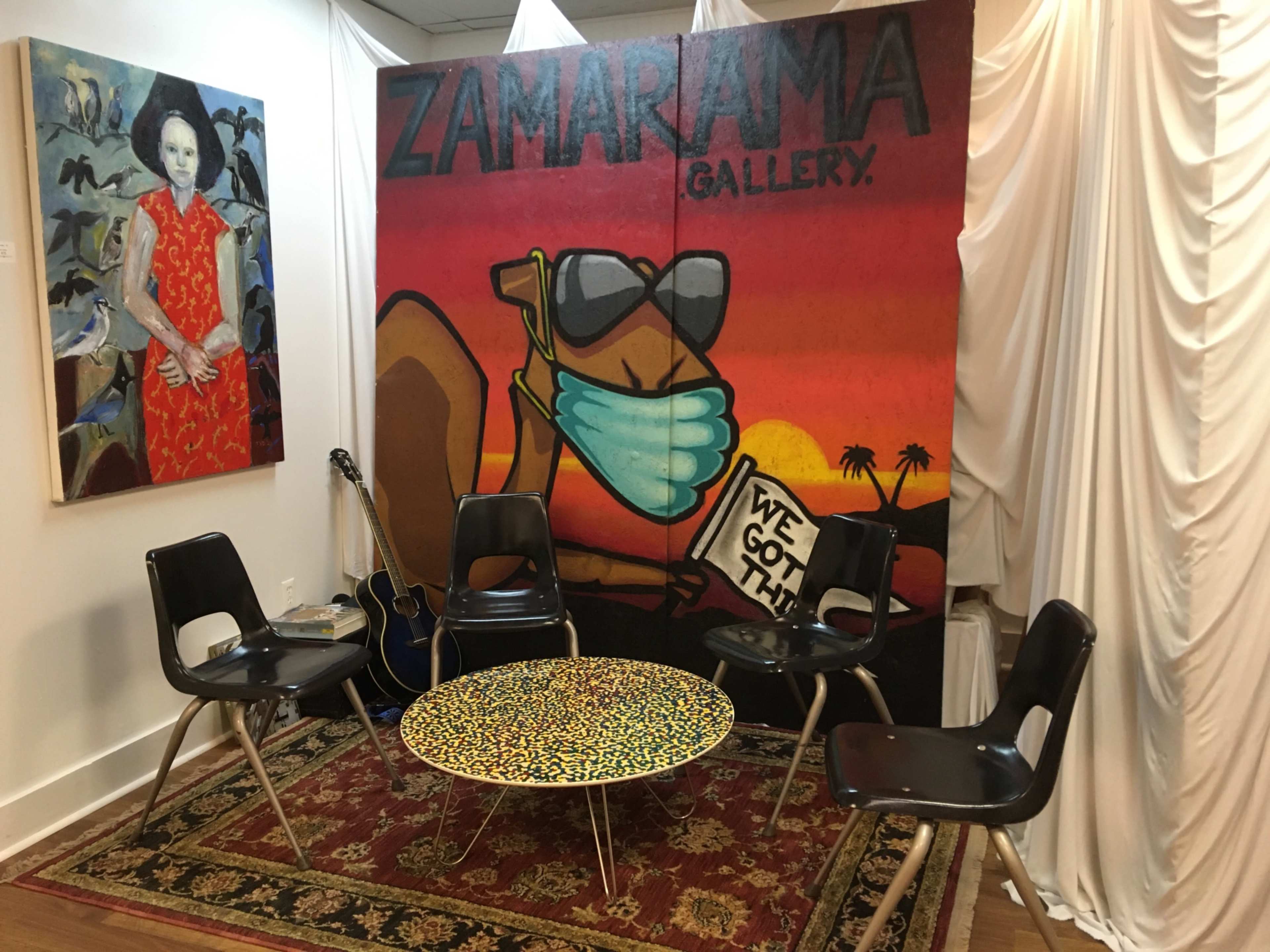 A small gallery space with a mural featuring a figure wearing a mask, surrounded by black chairs and a patterned table on a decorative rug.