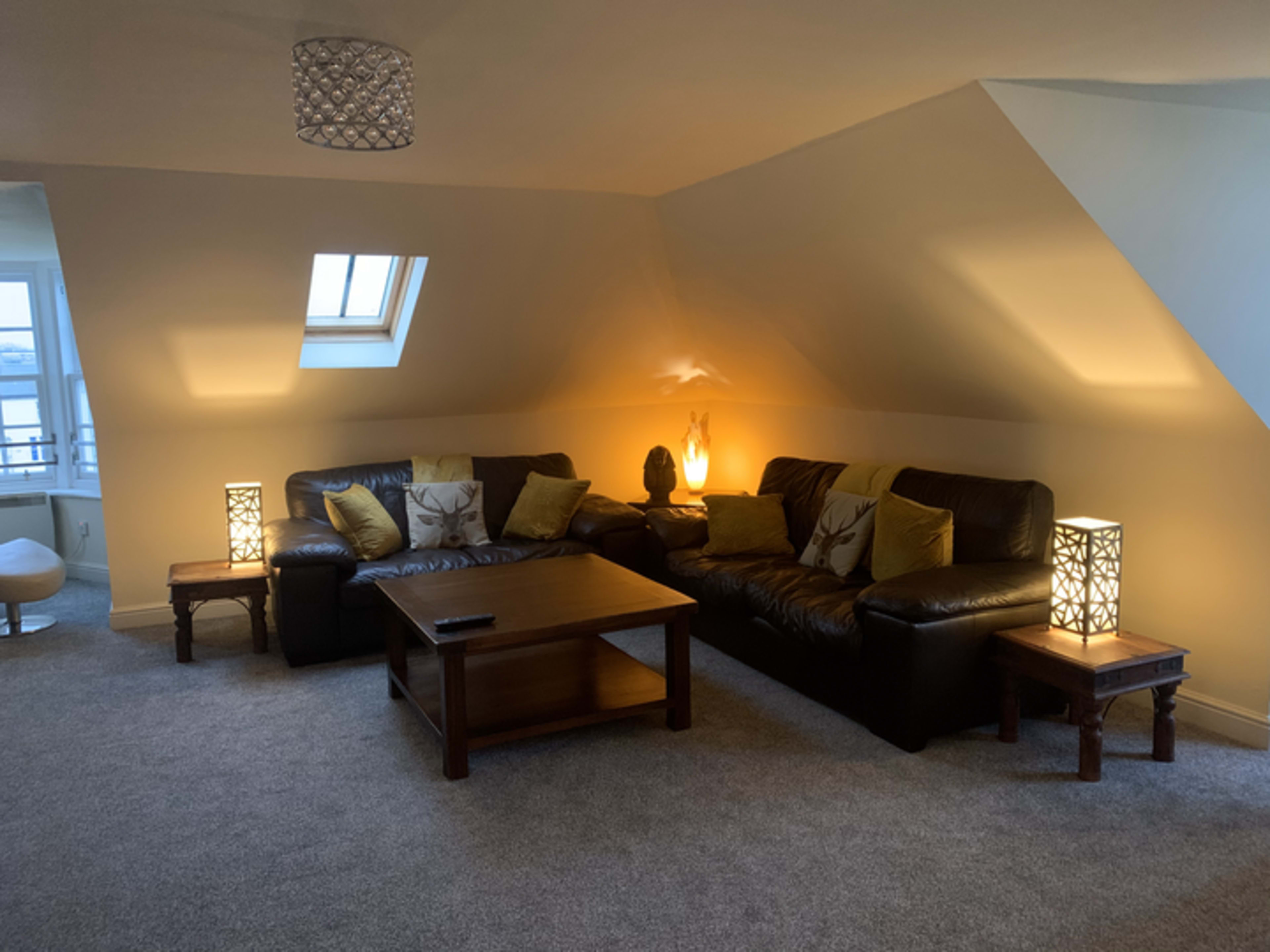 A cozy living room in an attic, featuring two dark leather sofas facing a wooden coffee table and illuminated by decorative lamps.