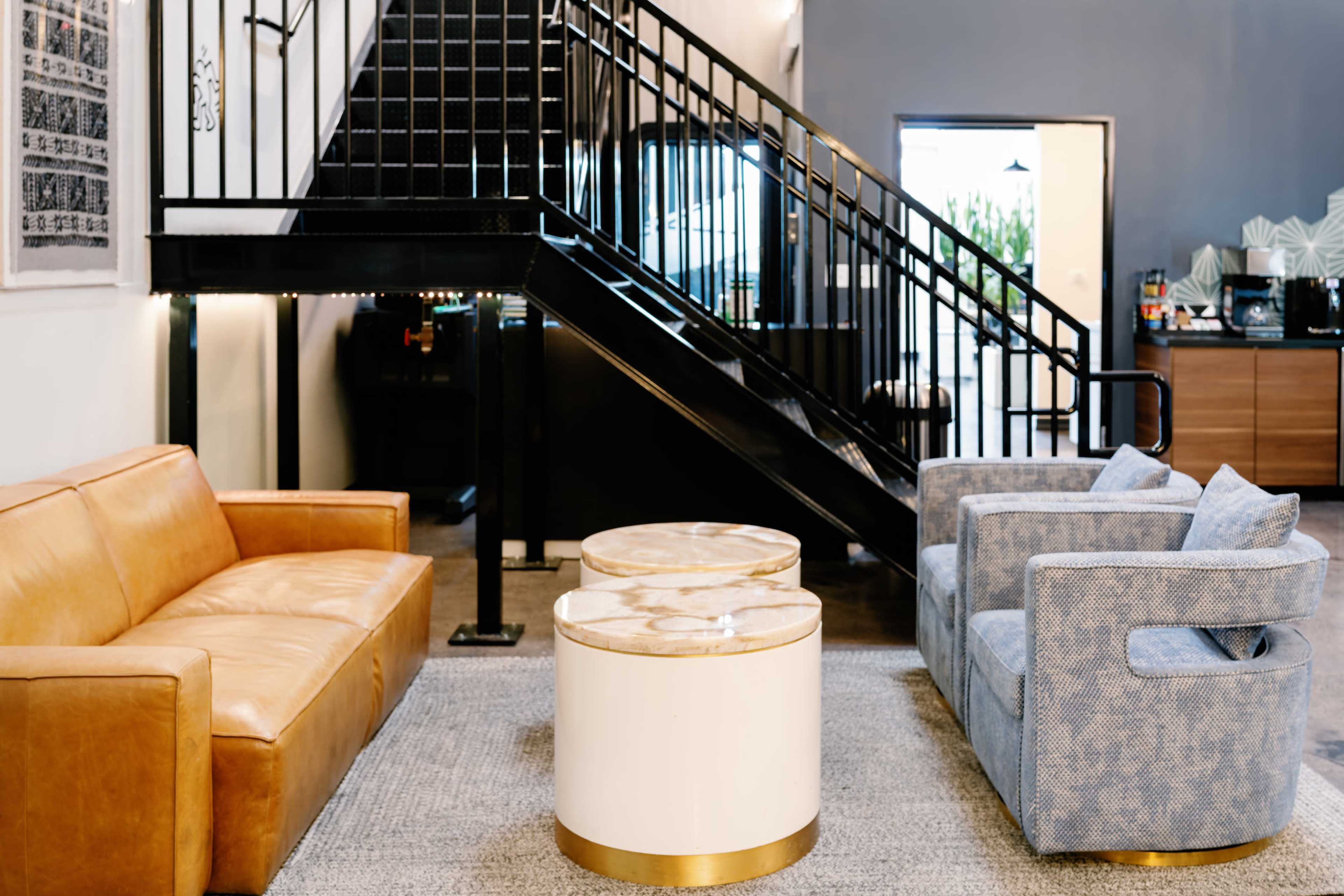 A brown leather sofa and a blue upholstered armchair are arranged across from two circular coffee tables on a gray rug under an industrial staircase.