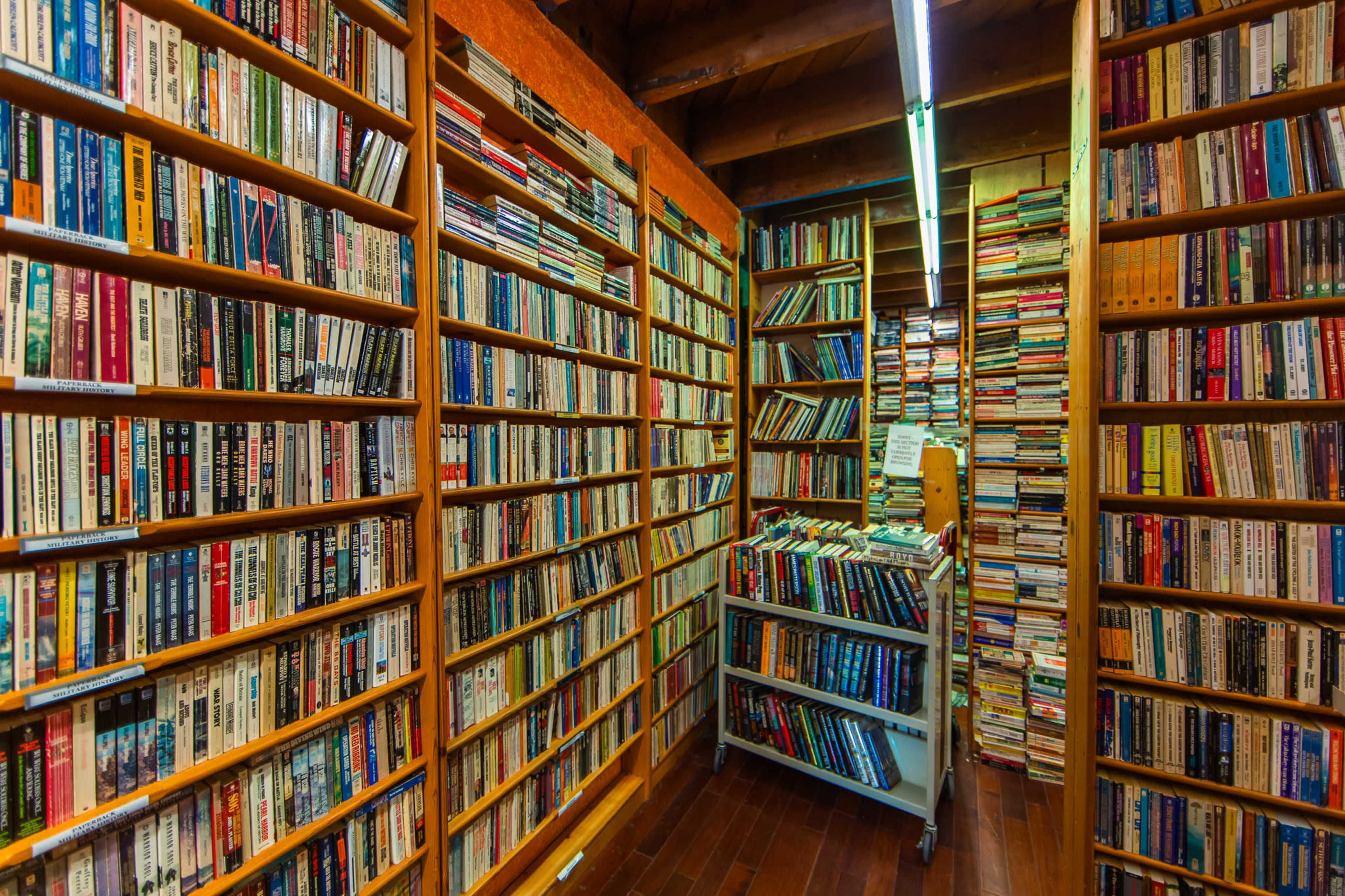 The image shows a narrow aisle flanked by tall bookshelves filled with numerous books, with a cart holding additional books in the center.