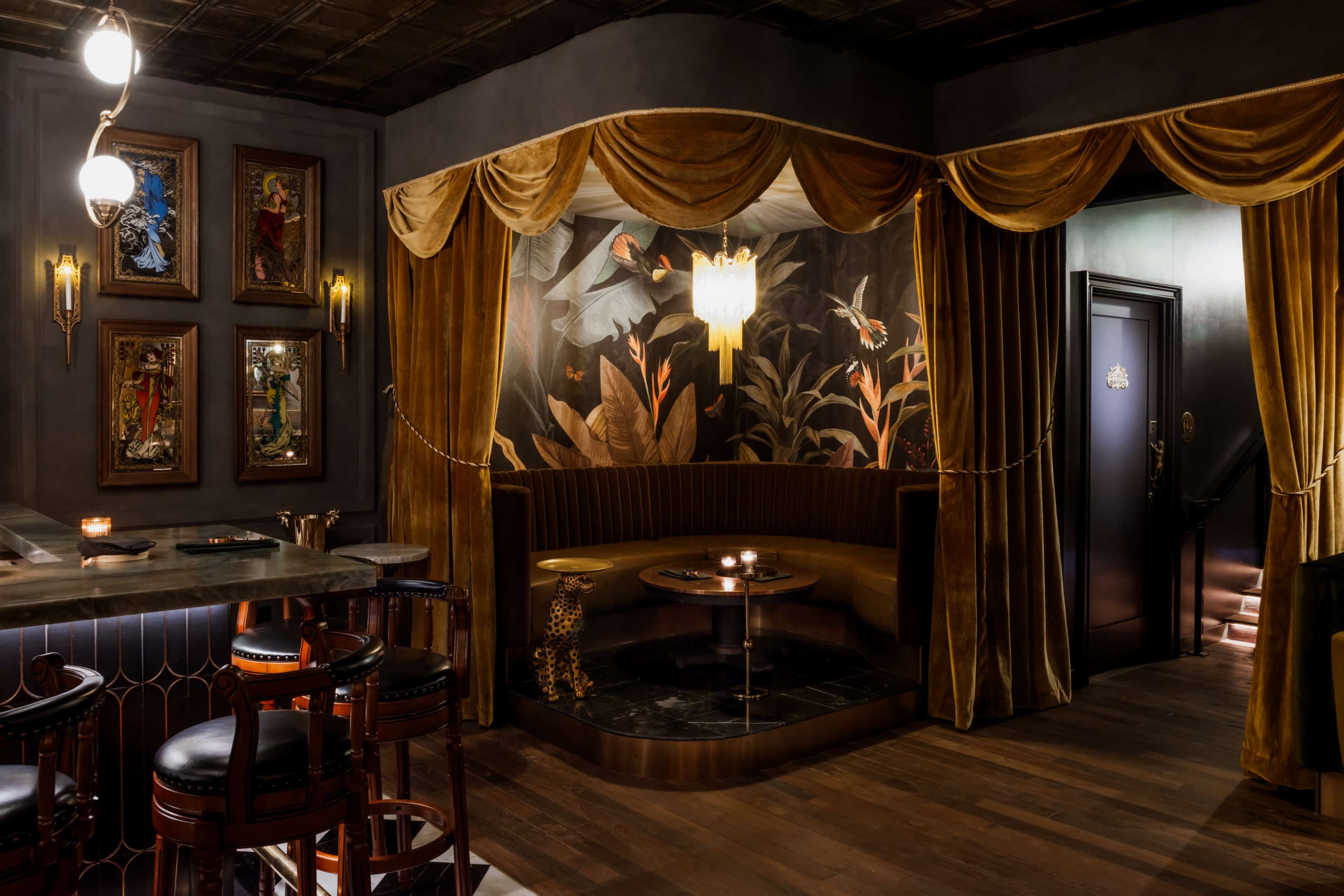 The image shows a dimly lit lounge area featuring rich, golden curtains, a plush velvet seating arrangement, and decorative artwork on the walls.