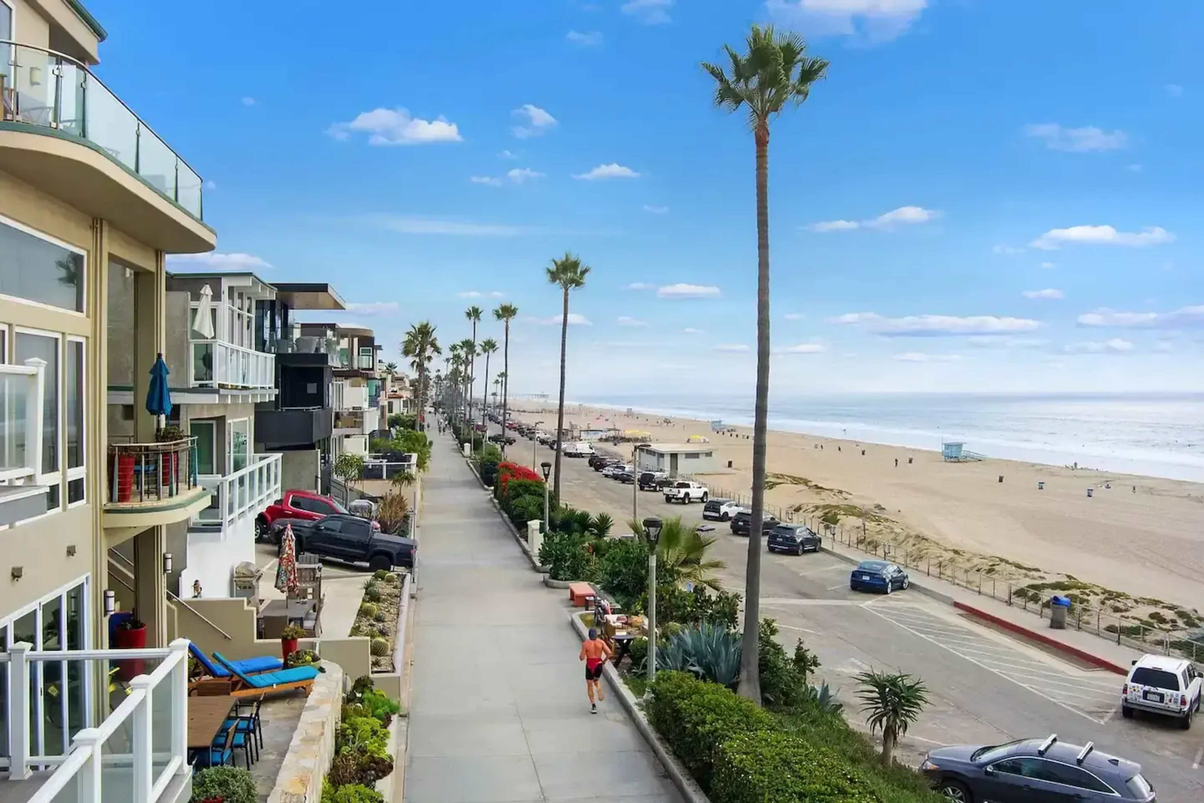 A paved walkway lined with palm trees runs parallel to a beach, with buildings and parked cars visible on one side.