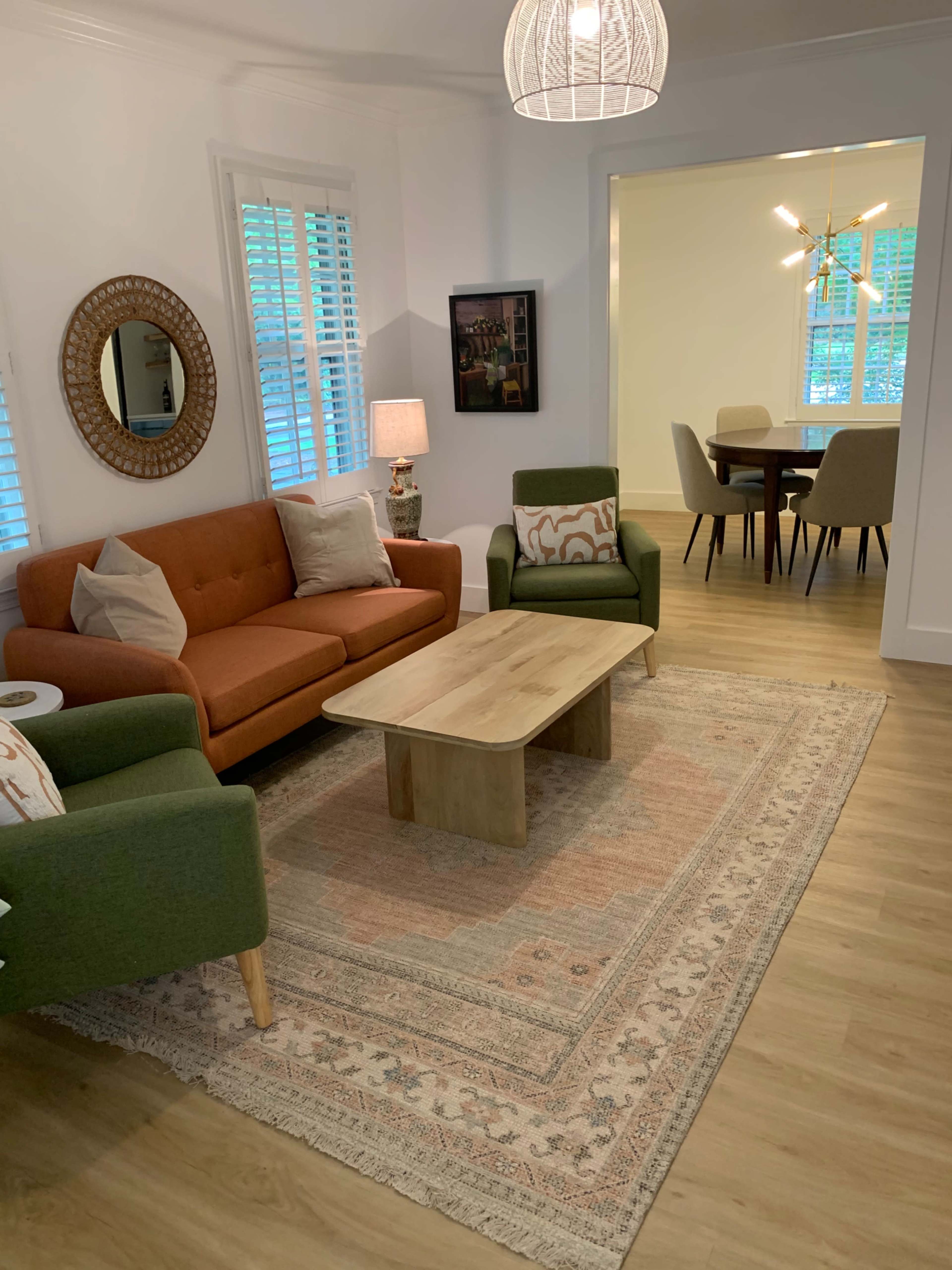 A cozy living room with a brown sofa, two green chairs, a wooden coffee table, and a patterned rug, illuminated by natural light through the window.