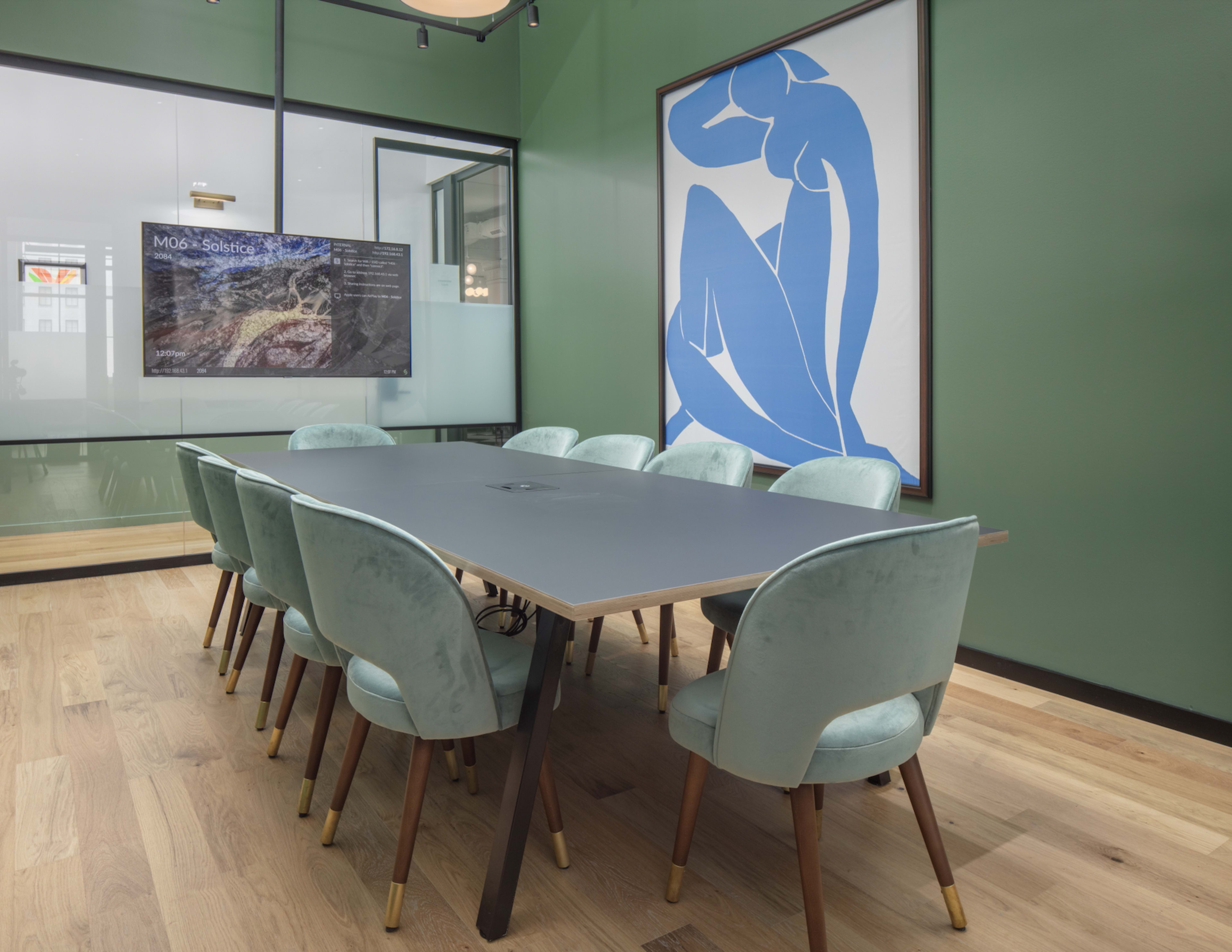 A conference room features a large table surrounded by upholstered chairs, with a wall-mounted screen and a large artwork.