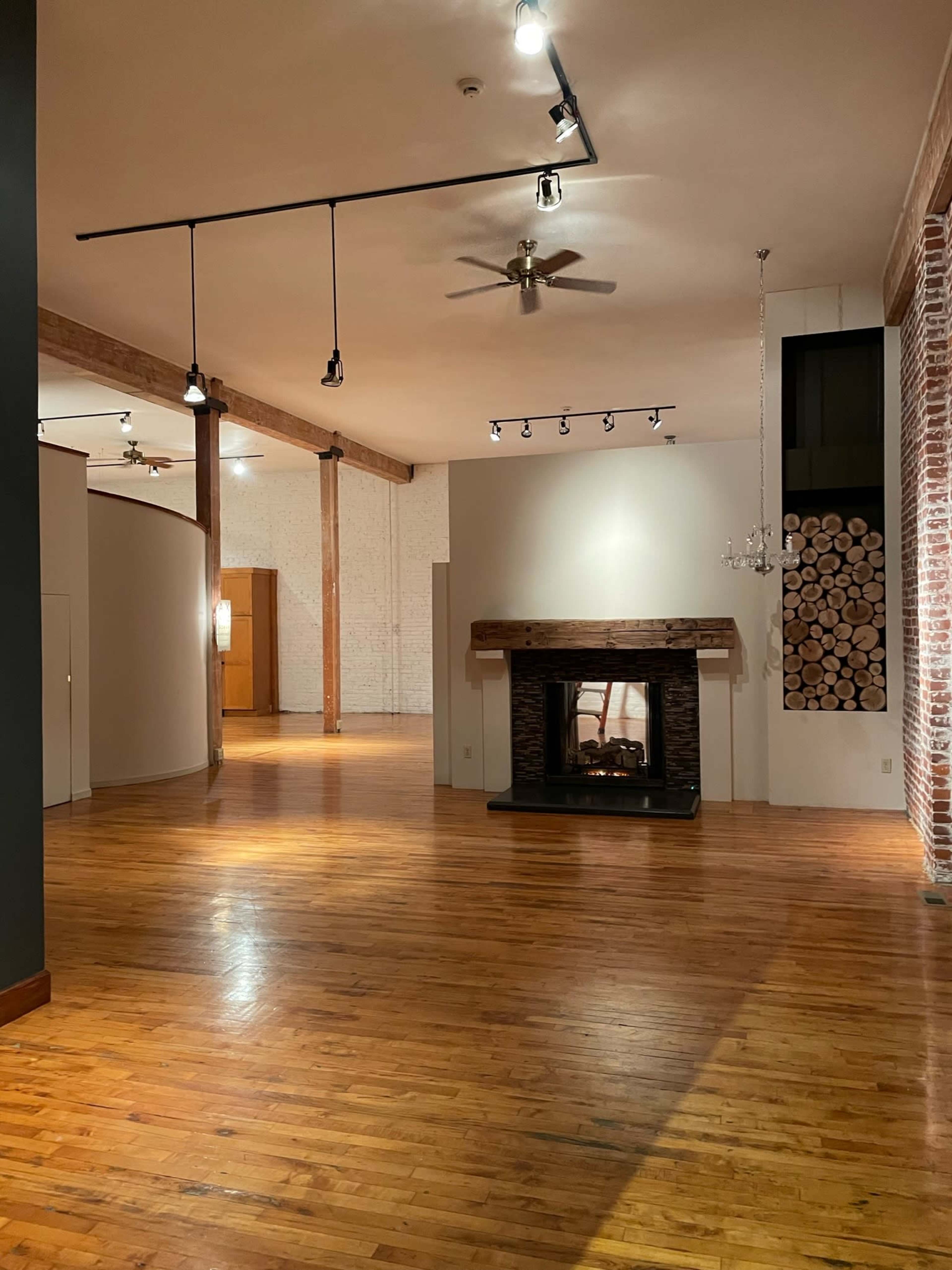 A spacious, well-lit room featuring wooden flooring, a fireplace with a stacked log accent wall, and high ceilings with exposed beams.