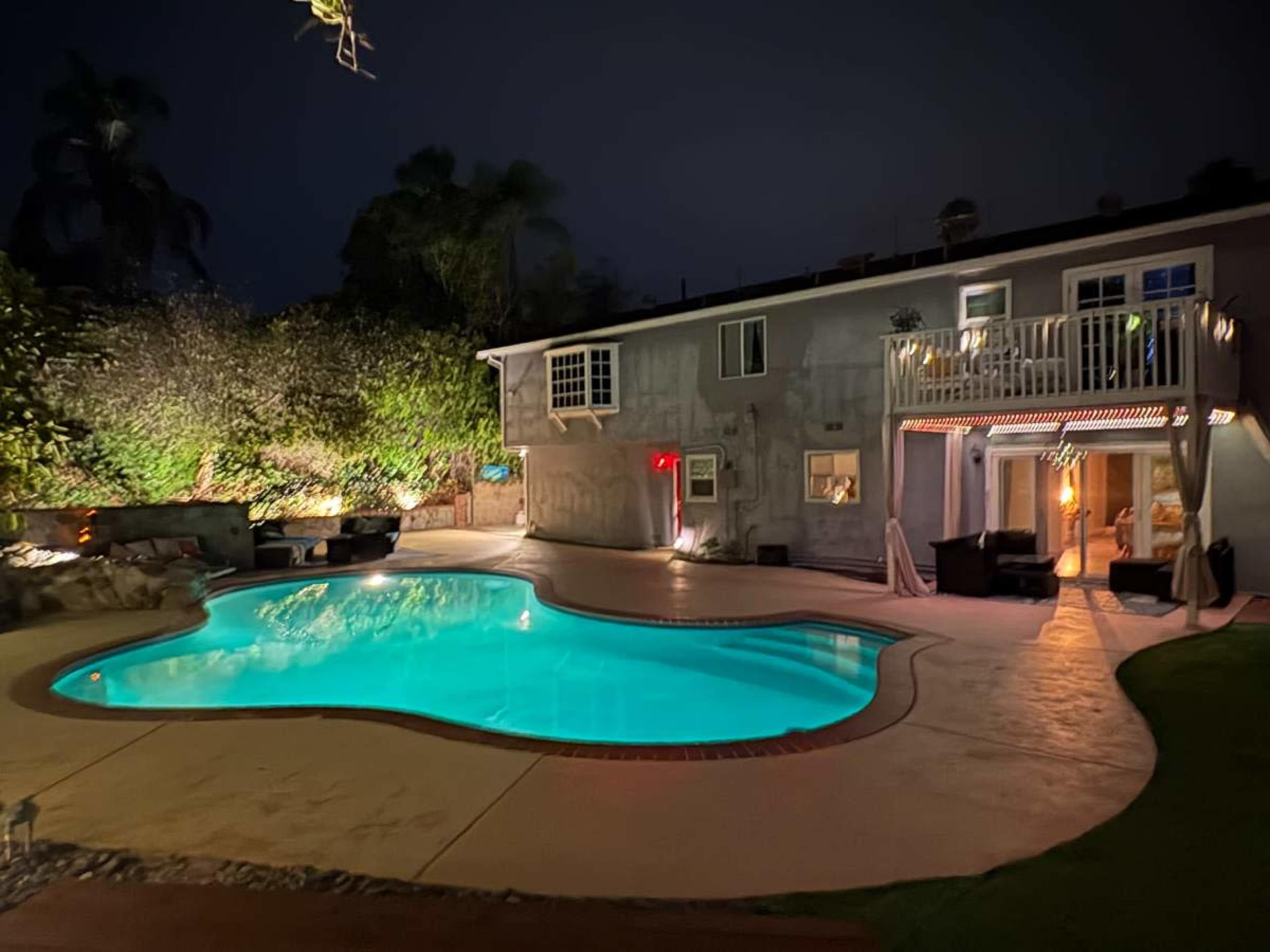The image shows a well-lit swimming pool in a backyard at night, surrounded by trees and a two-story house with an illuminated balcony.