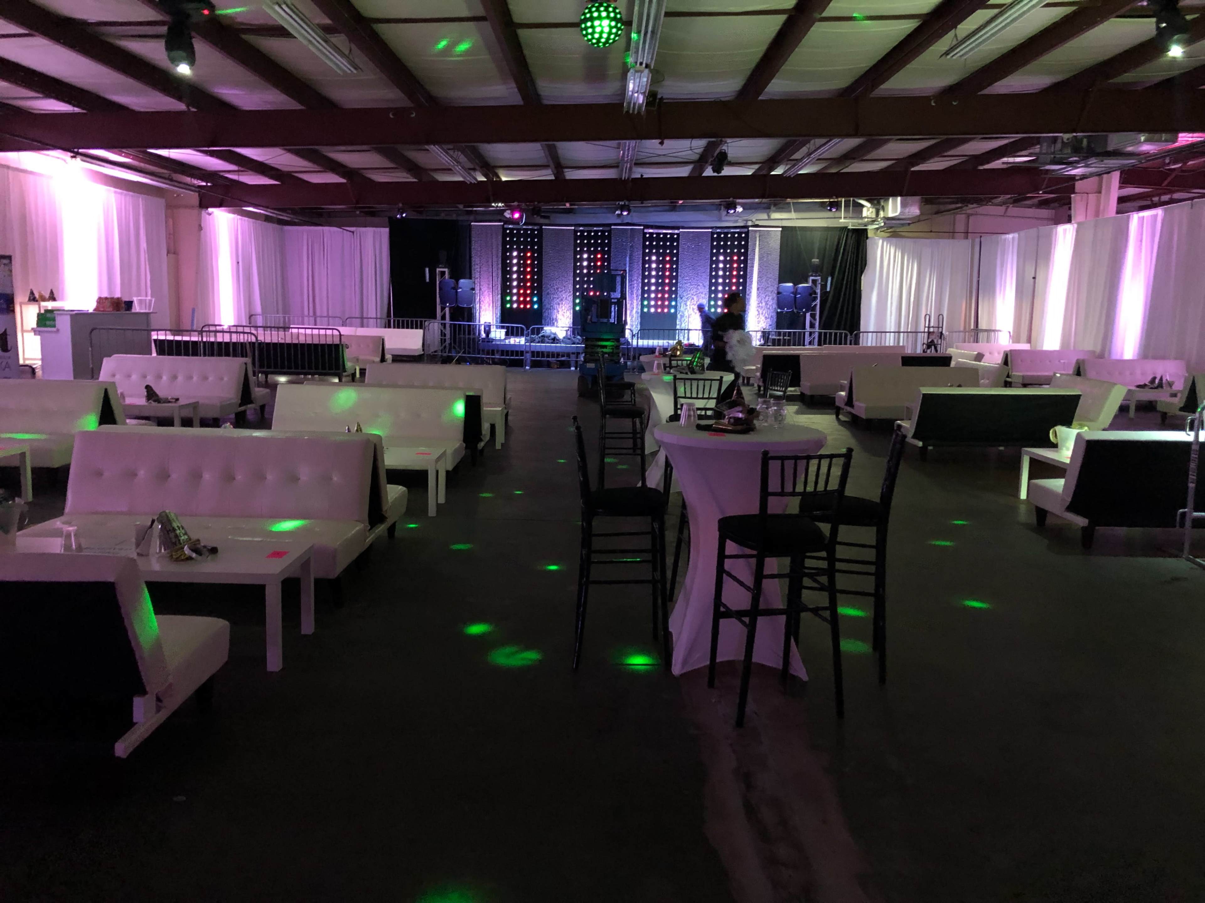 The image shows a spacious event venue with white seating, tables, and stage lighting set up for a gathering.