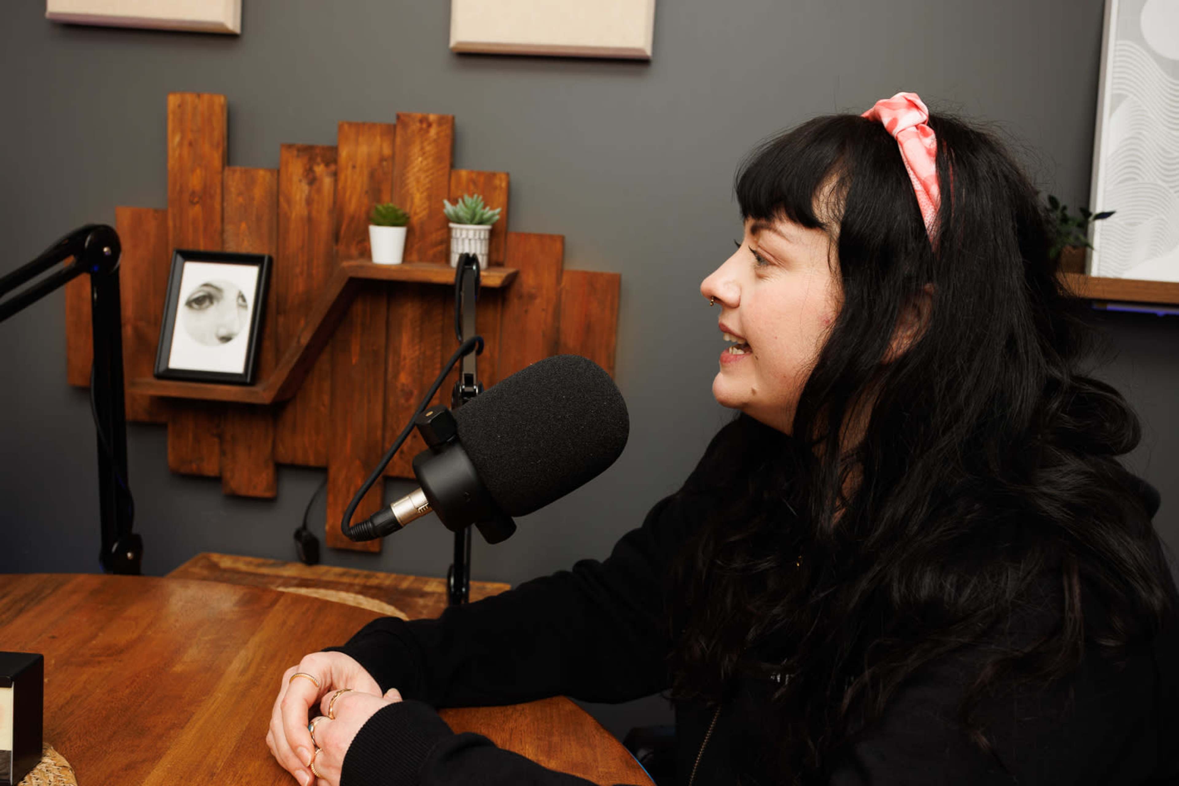 A woman with dark hair and a pink headband is sitting at a wooden table, speaking into a microphone in a room with modern decor.