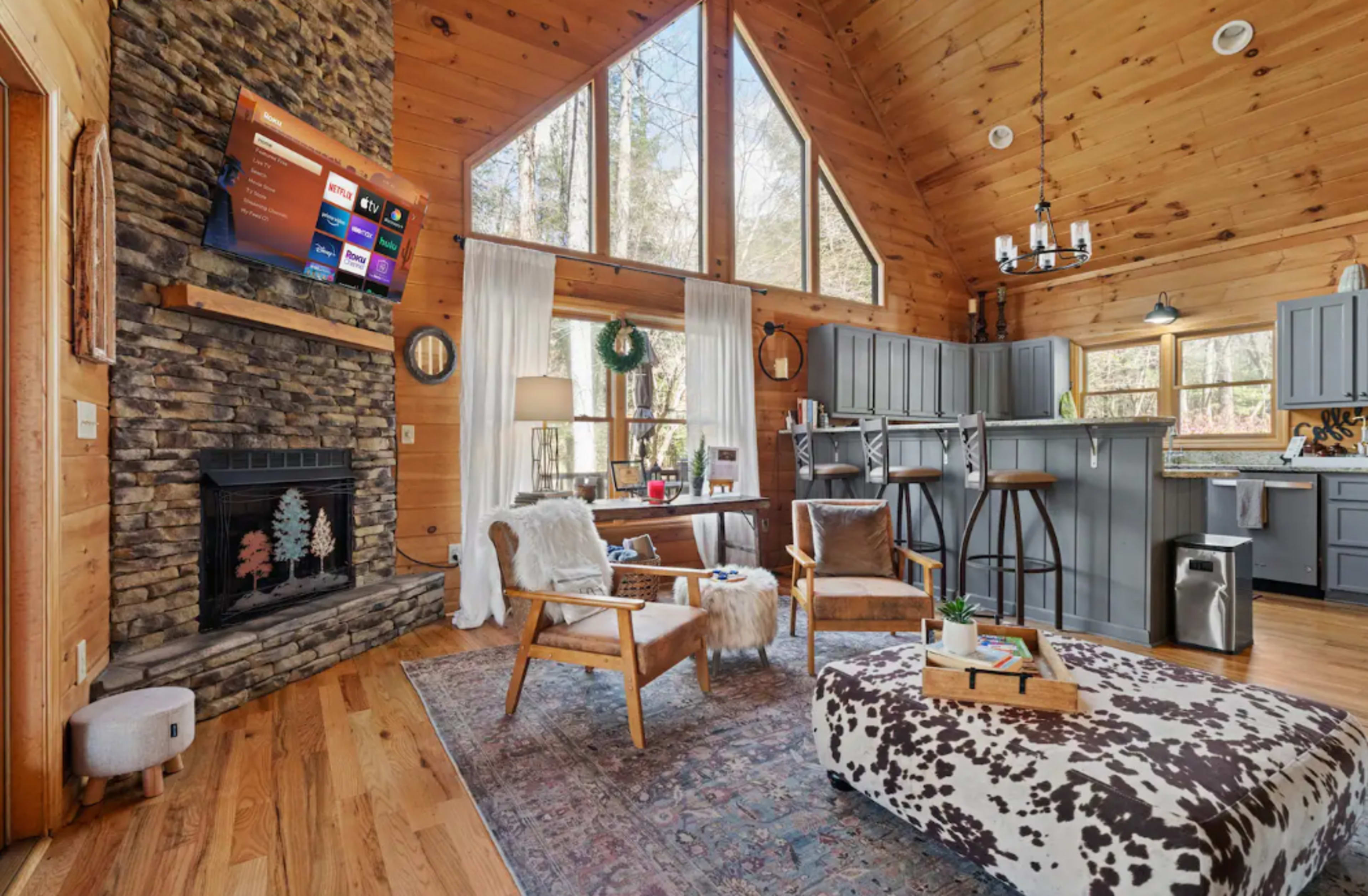 A cozy cabin interior features a stone fireplace, wooden furniture, and large windows, allowing natural light to fill the space.