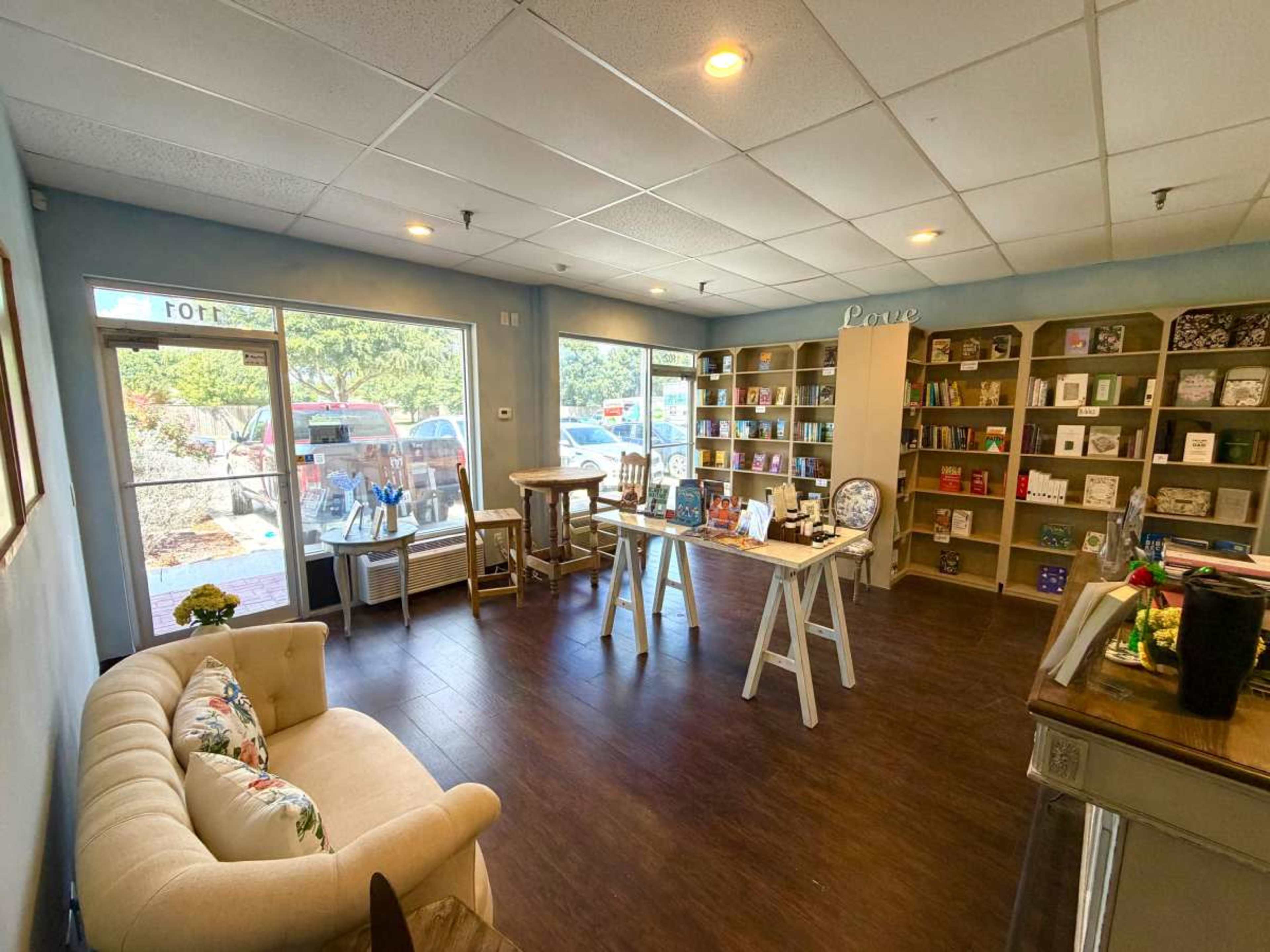 The image shows a neatly arranged interior of a store with shelves filled with various products, a beige couch, and a large window allowing natural light to enter.
