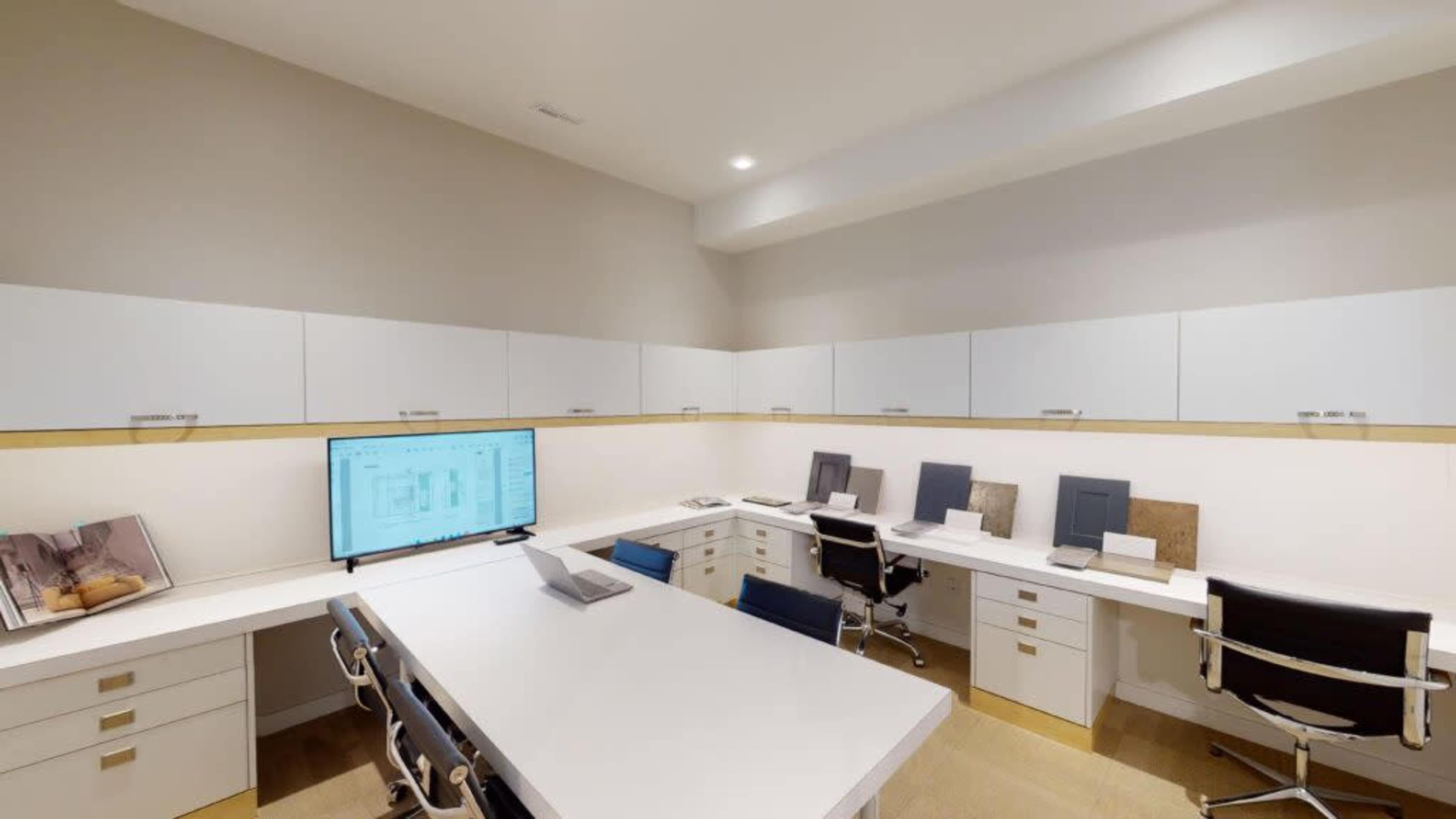 A modern office space featuring white desks, sleek chairs, and a large monitor displaying plans on the wall.