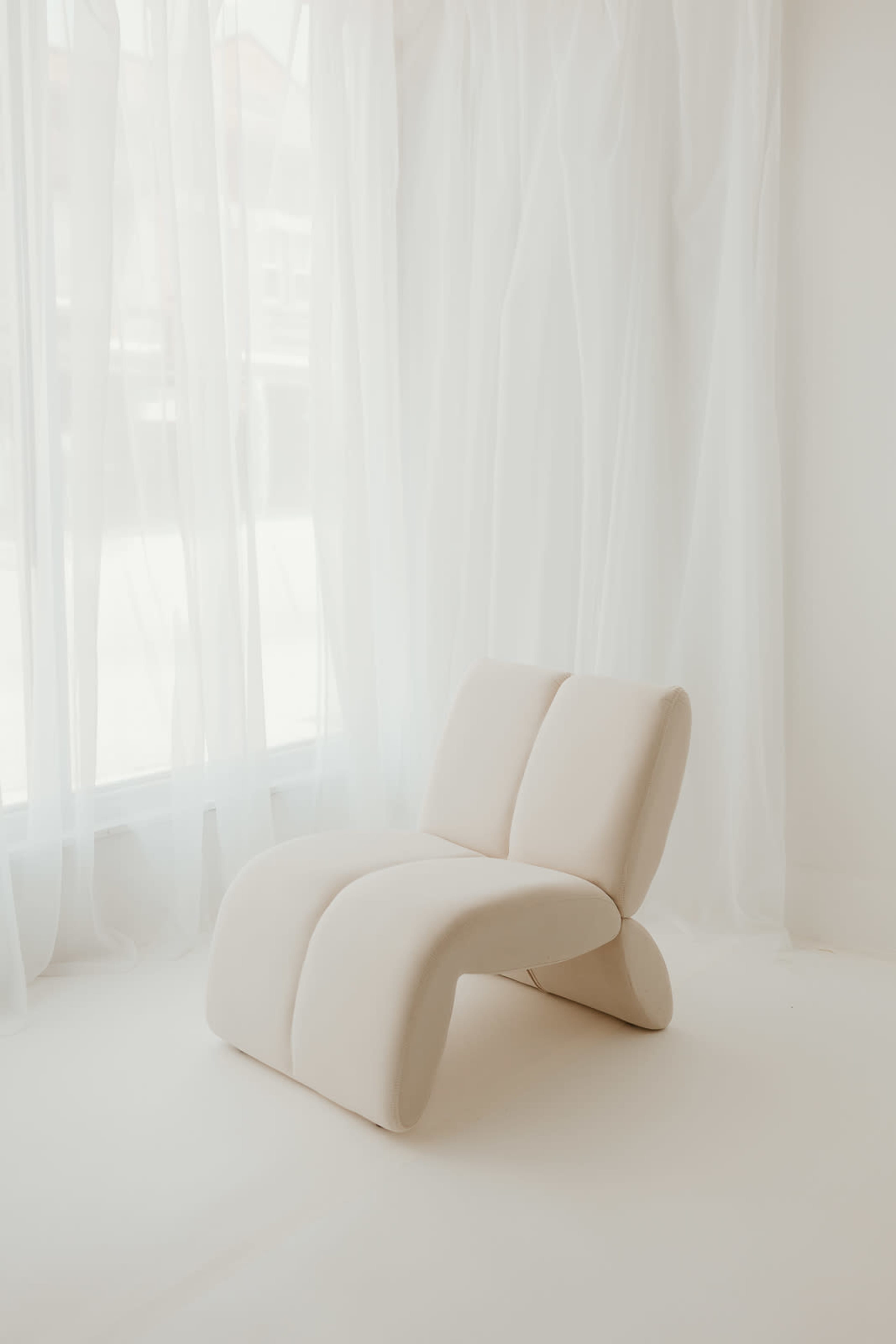 A minimalist, white lounge chair is positioned near a large window draped with sheer curtains.