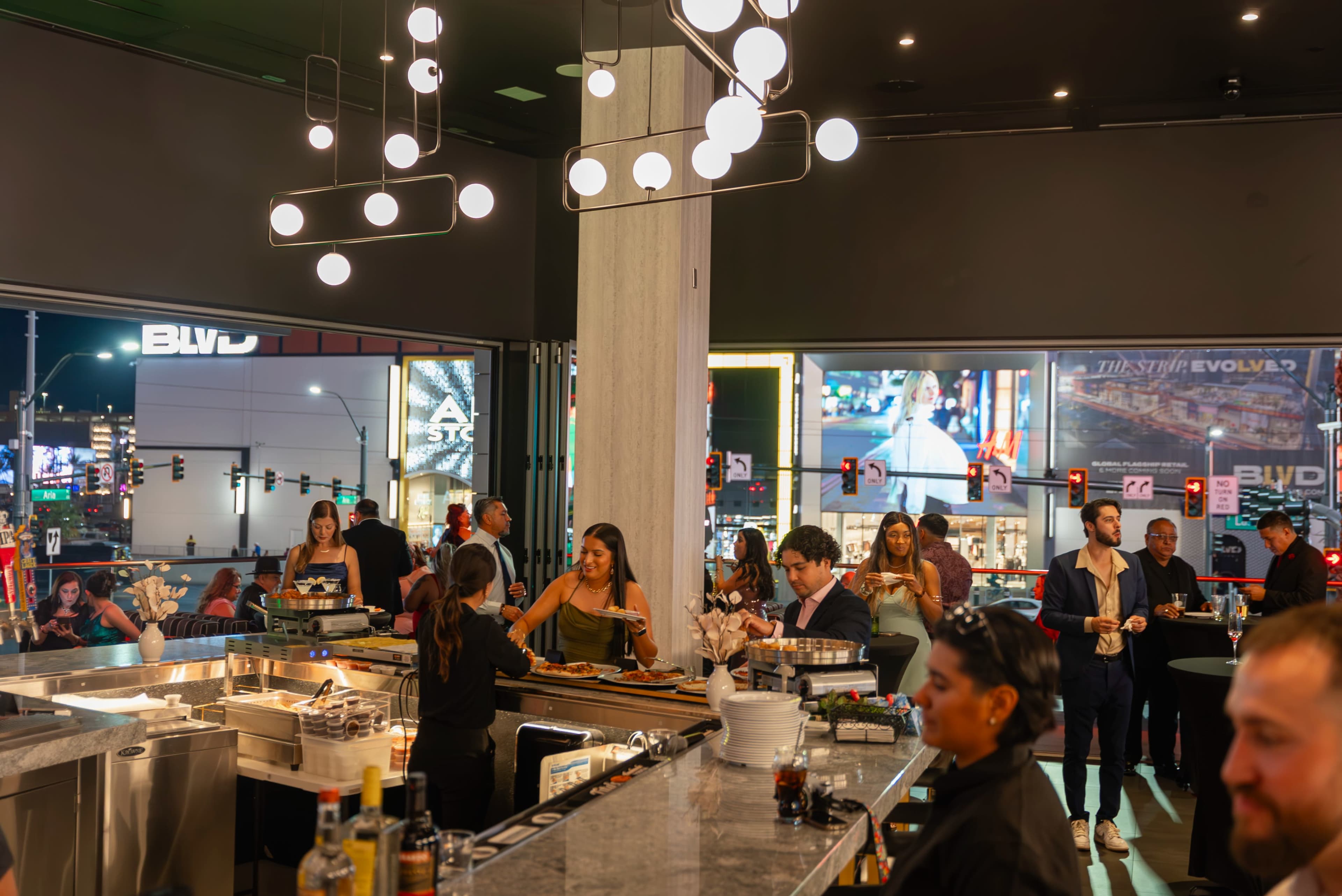 A bustling bar scene features patrons socializing and dining while a vibrant cityscape glows through the large windows.
