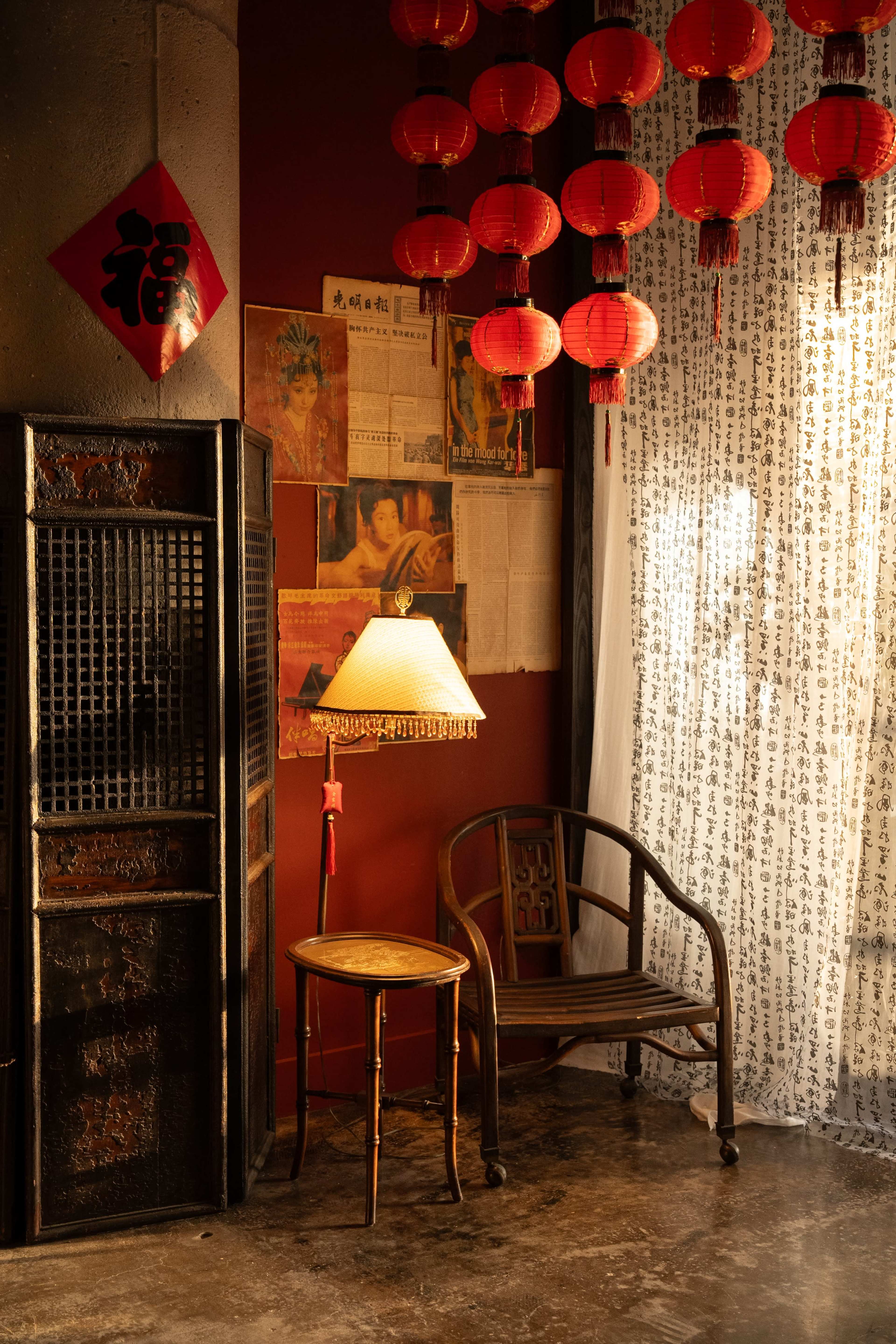A cozy corner of a room adorned with red lanterns, a lamp on a small round table, and a wooden chair beside a wall featuring Chinese calligraphy and vintage posters.