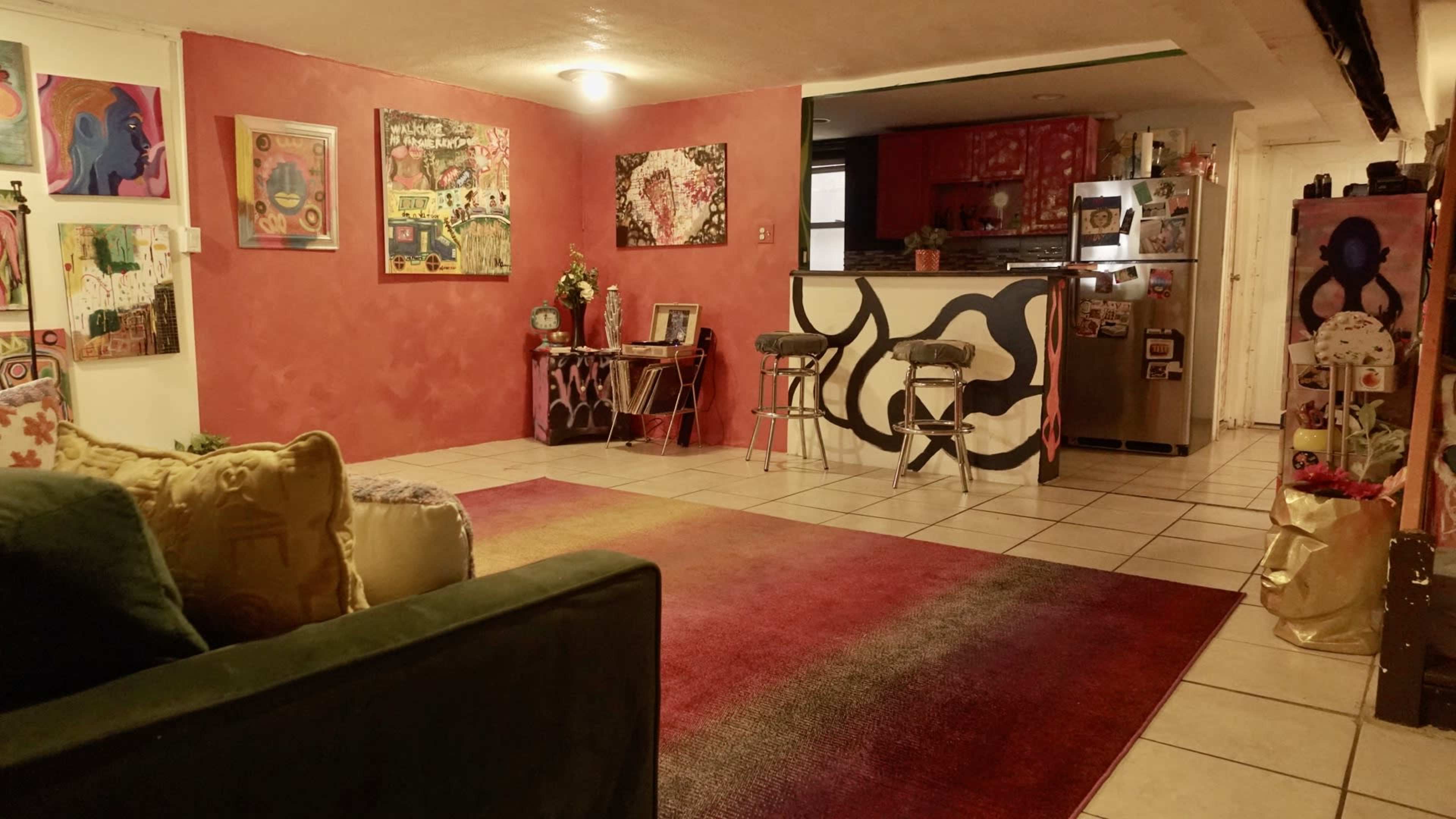 A brightly colored living room features a large area rug, eclectic artwork on the walls, and a kitchen visible in the background with a bar counter and stools.