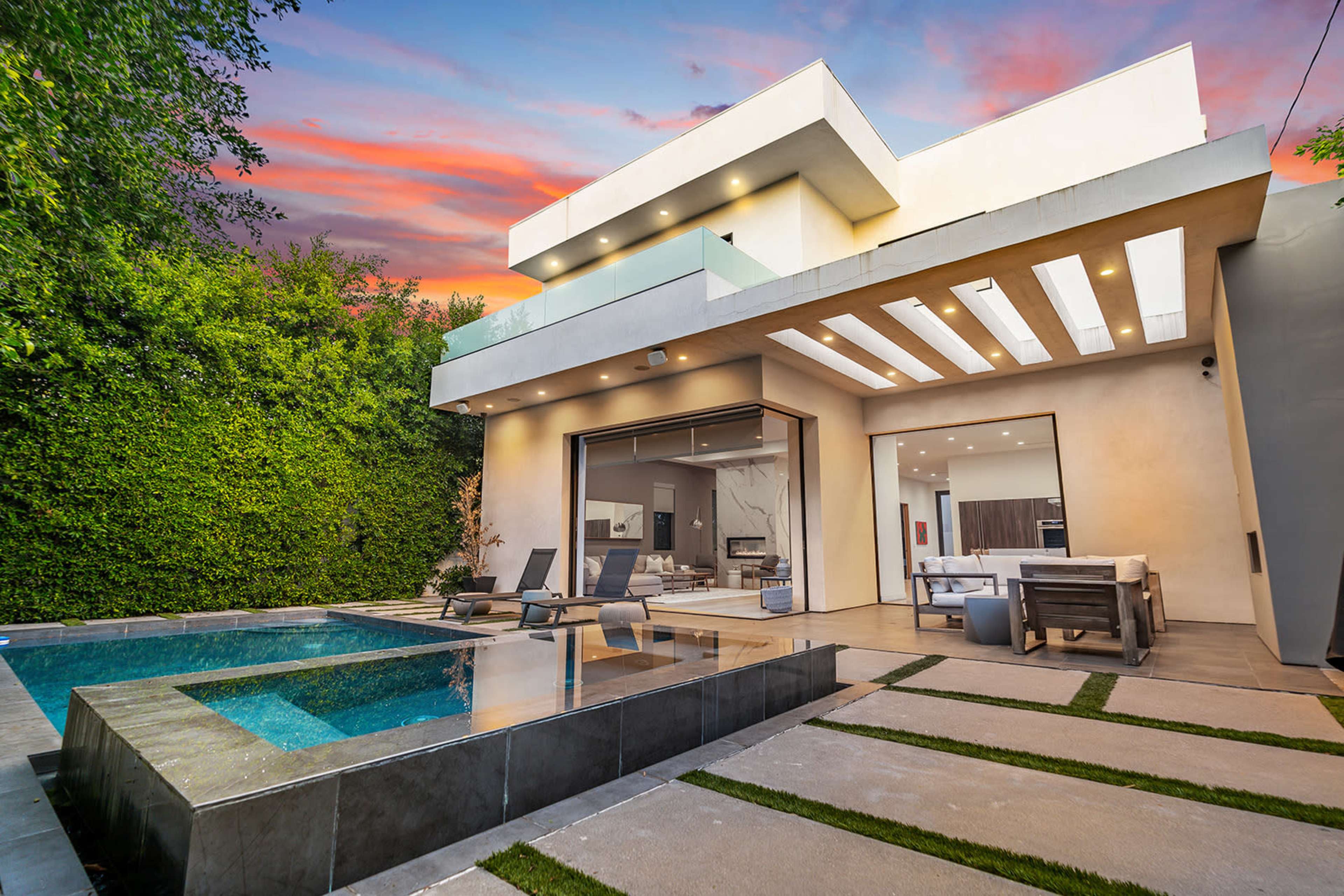 A modern house with a pool is set against a colorful sunset and surrounded by greenery.