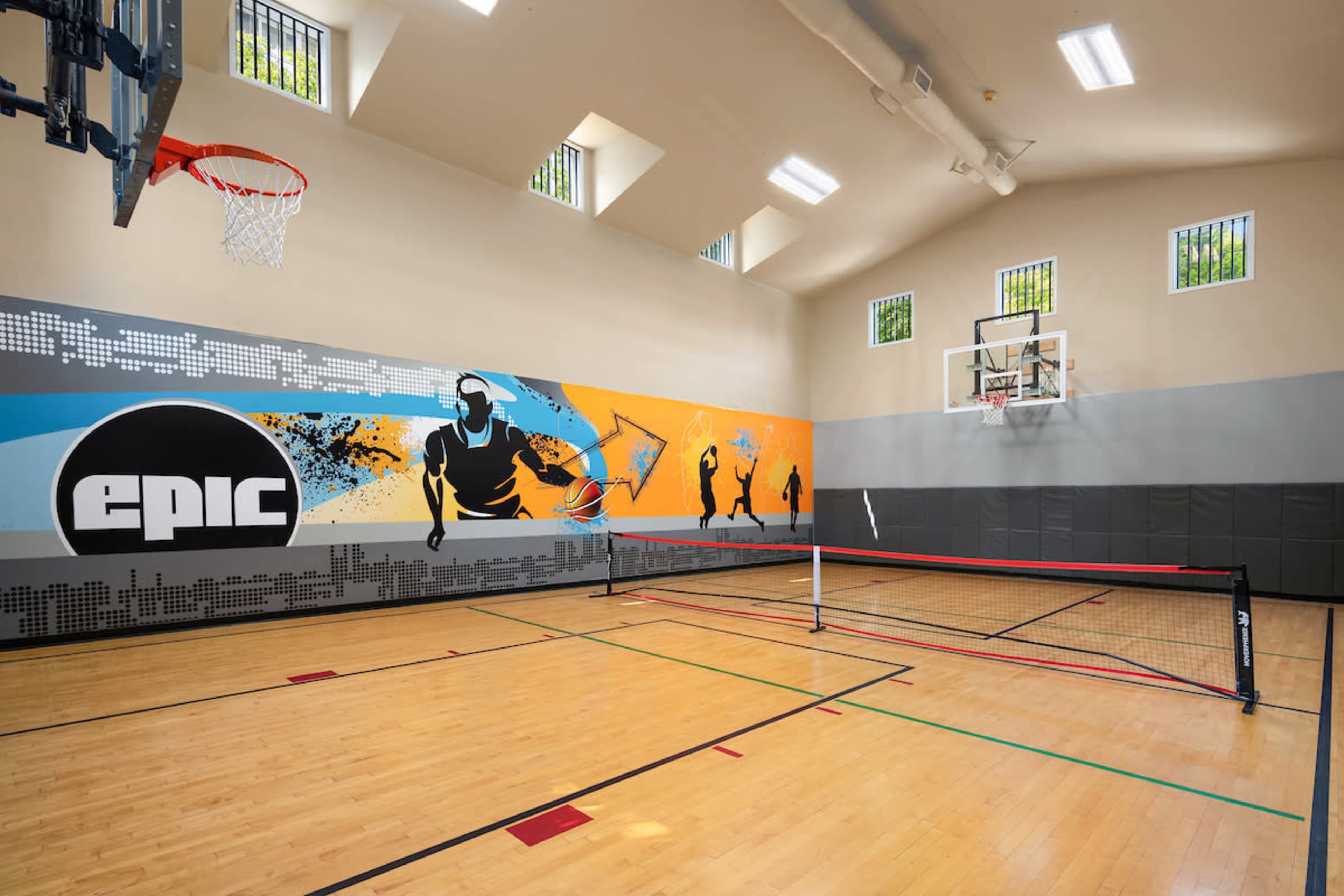 The image shows an indoor sports facility featuring a basketball hoop on the wall and a volleyball net set up on a wooden court, with colorful murals depicting athletic figures.
