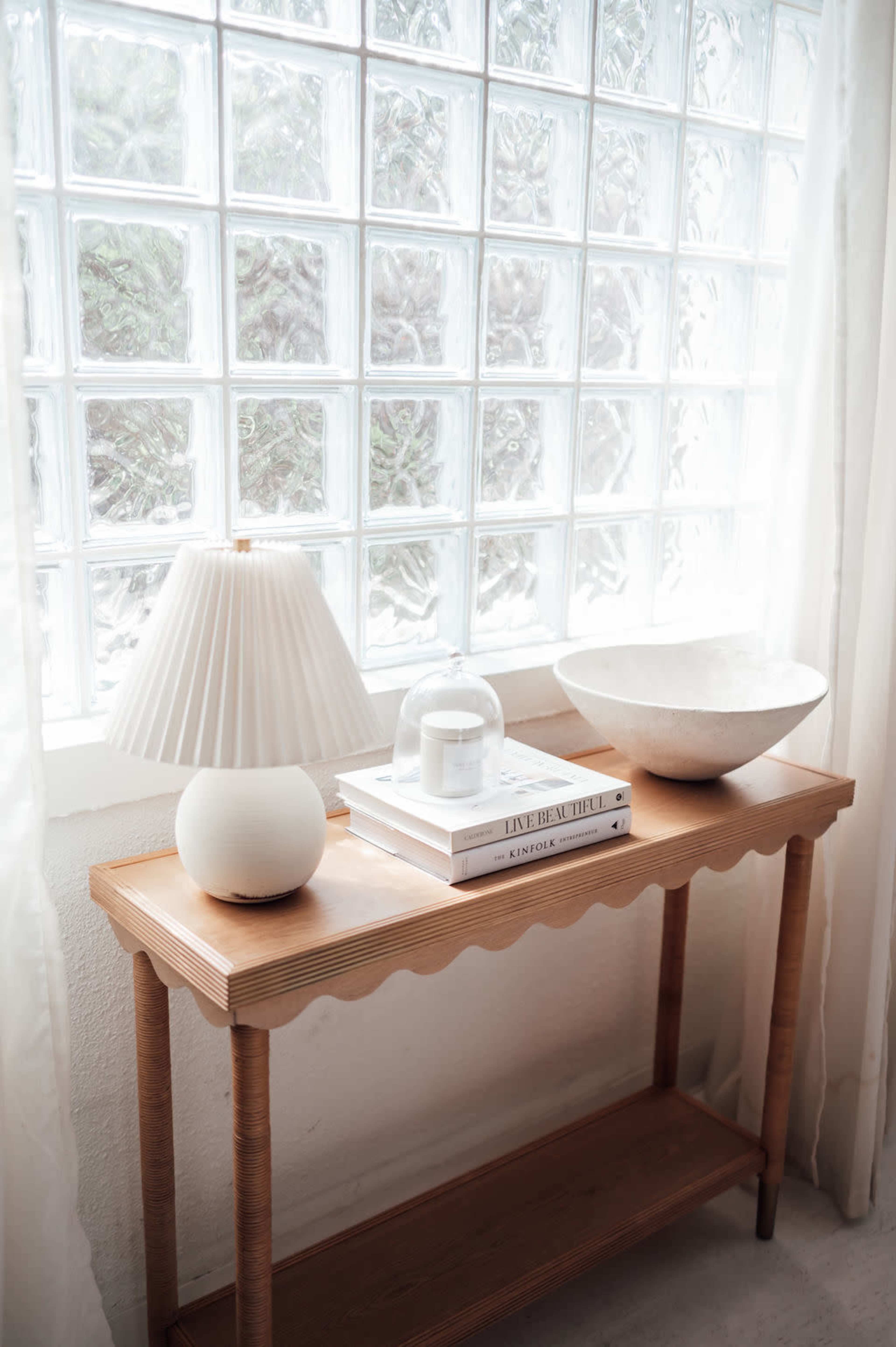 A wooden console table with a scalloped edge holds a lamp, a decorative bowl, and a stack of books, positioned near a glass block window.