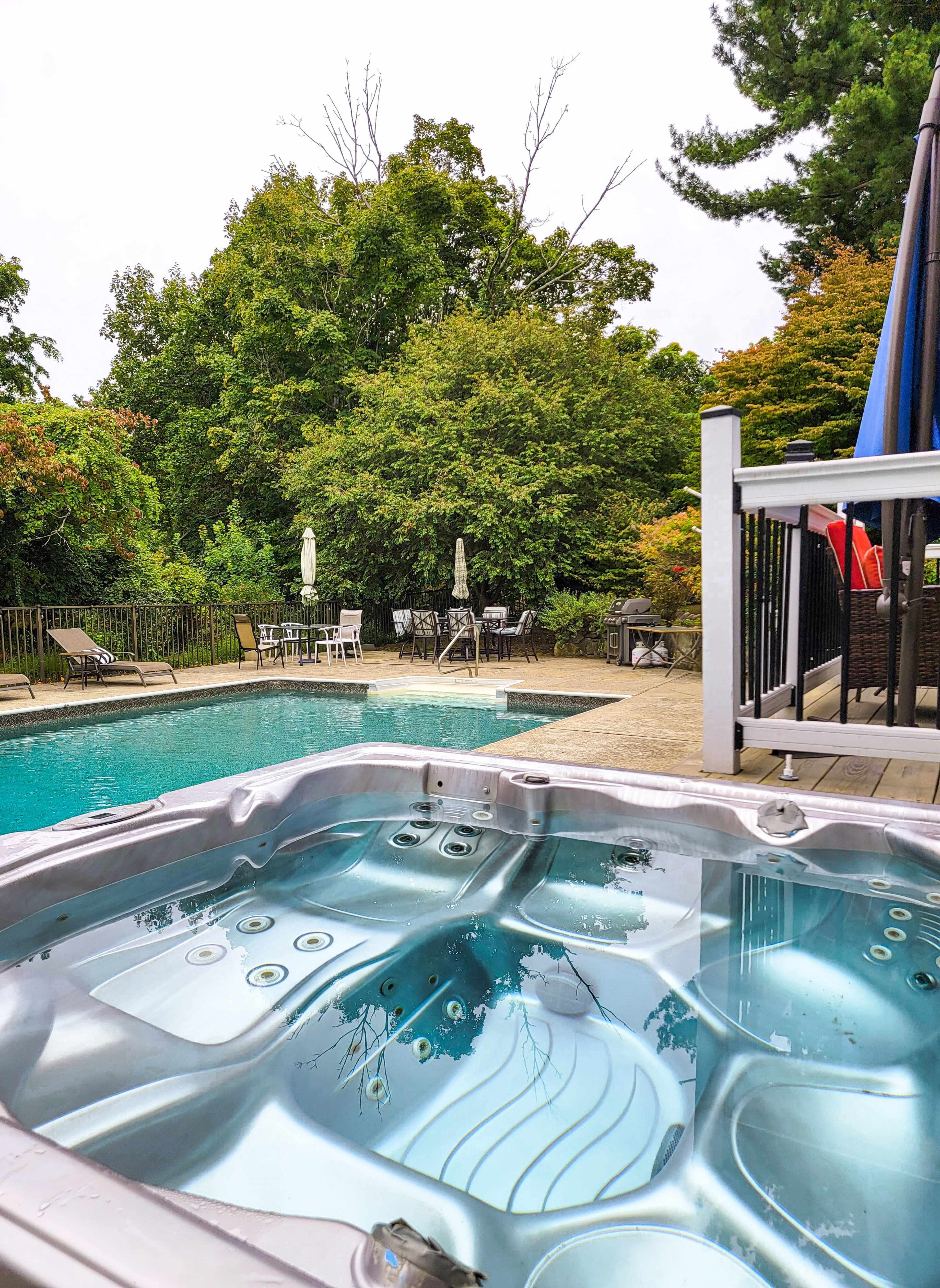 A hot tub is situated in the foreground, overlooking a pool area surrounded by trees and patio furniture.