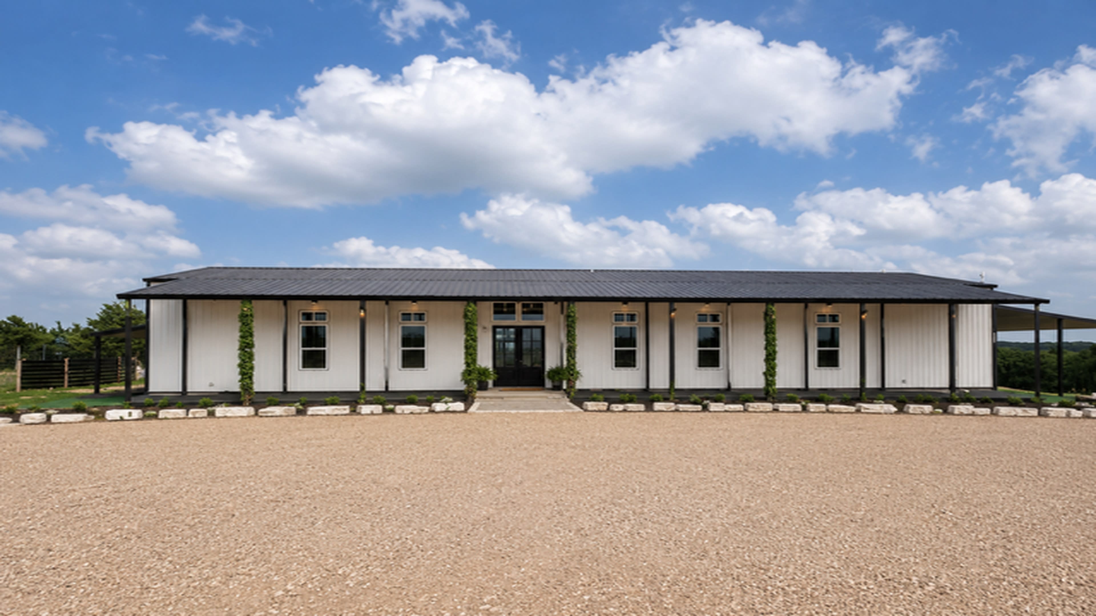 The image shows a modern, single-story building with a white exterior, a dark roof, and large windows, set against a backdrop of a blue sky with fluffy clouds.