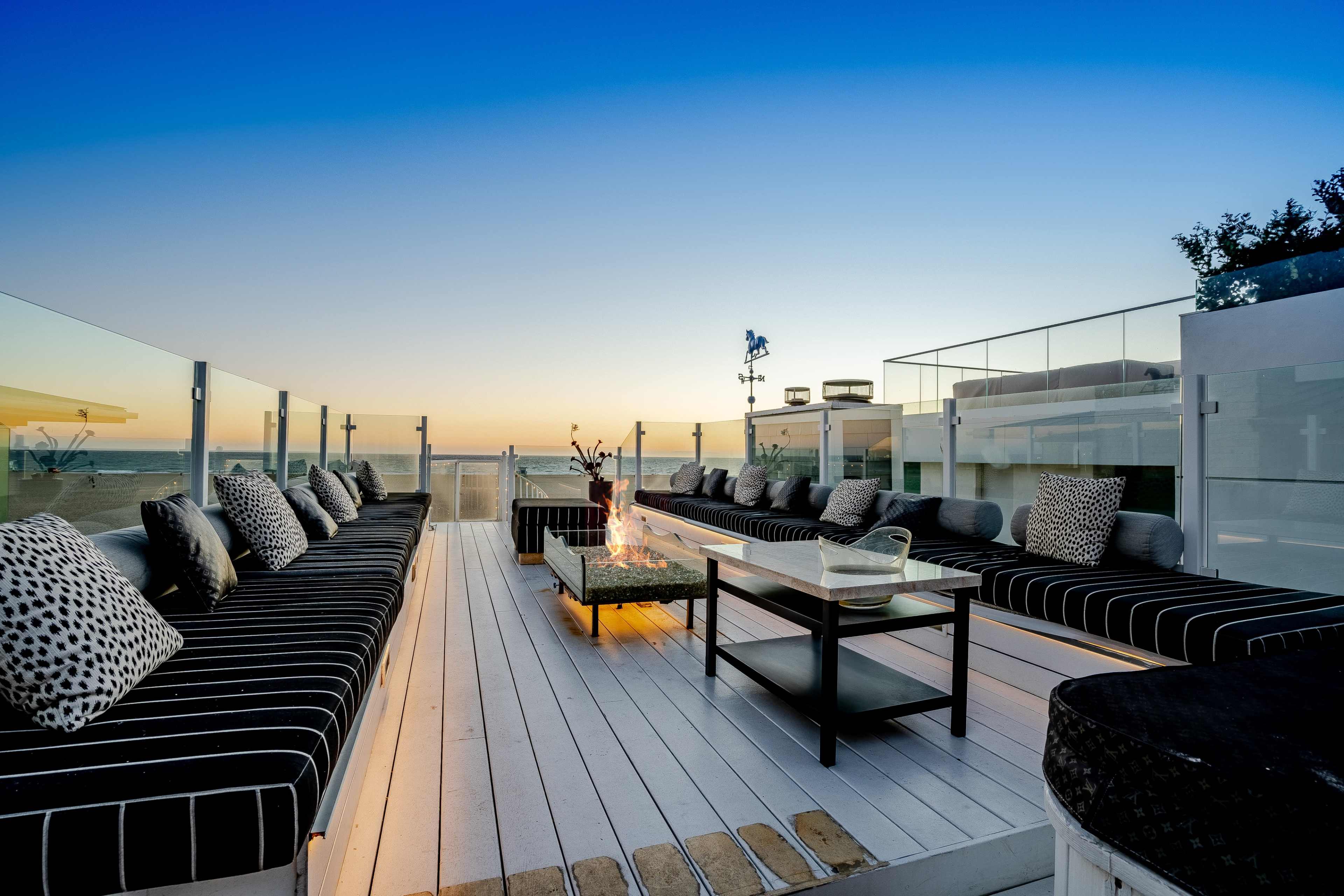 A rooftop terrace features a long seating area with striped cushions, a central coffee table, and a fire pit, all set against a sunset view over the ocean.