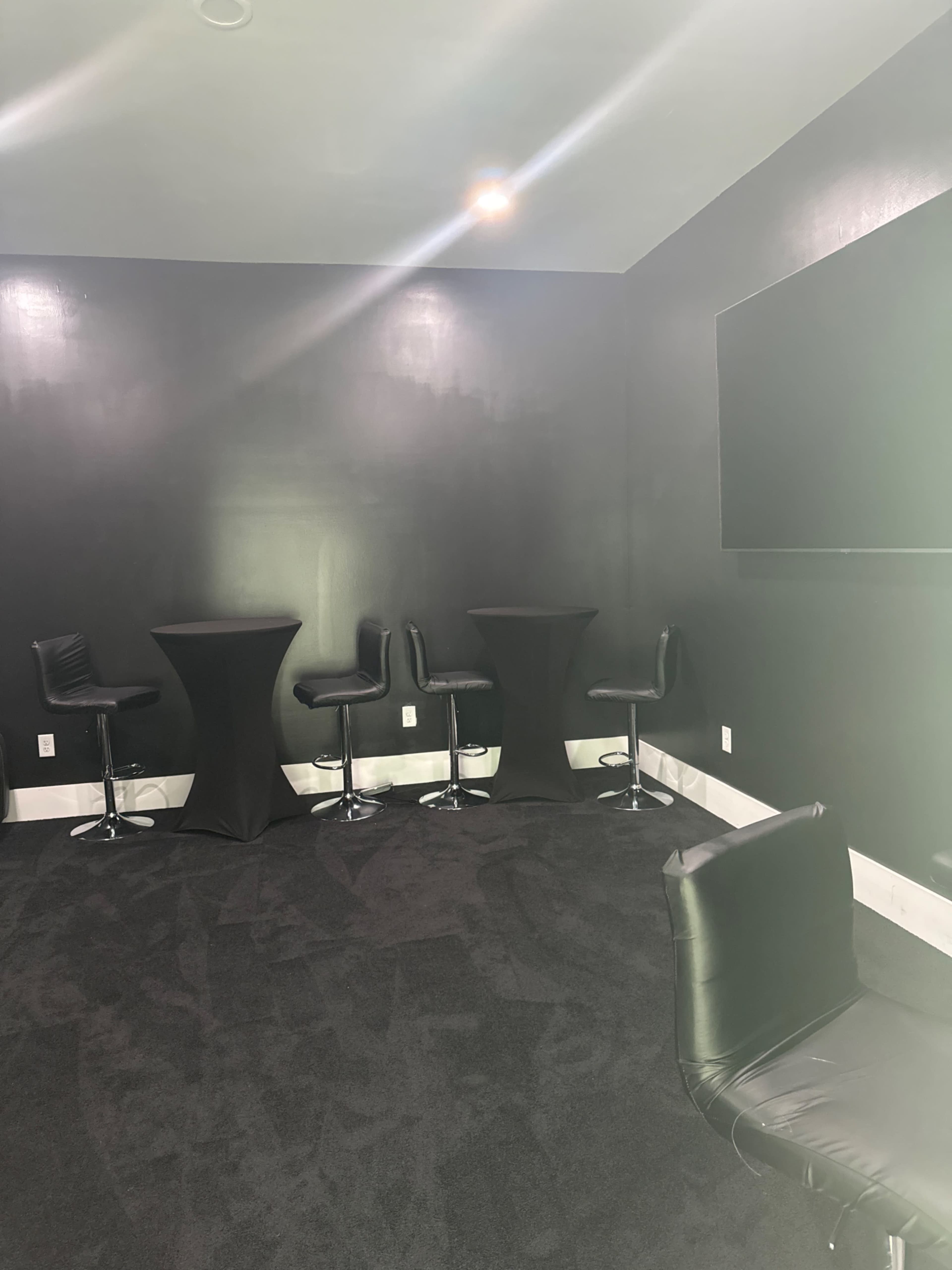 The image shows a small room with black walls, featuring two tall black tables and several black bar stools.