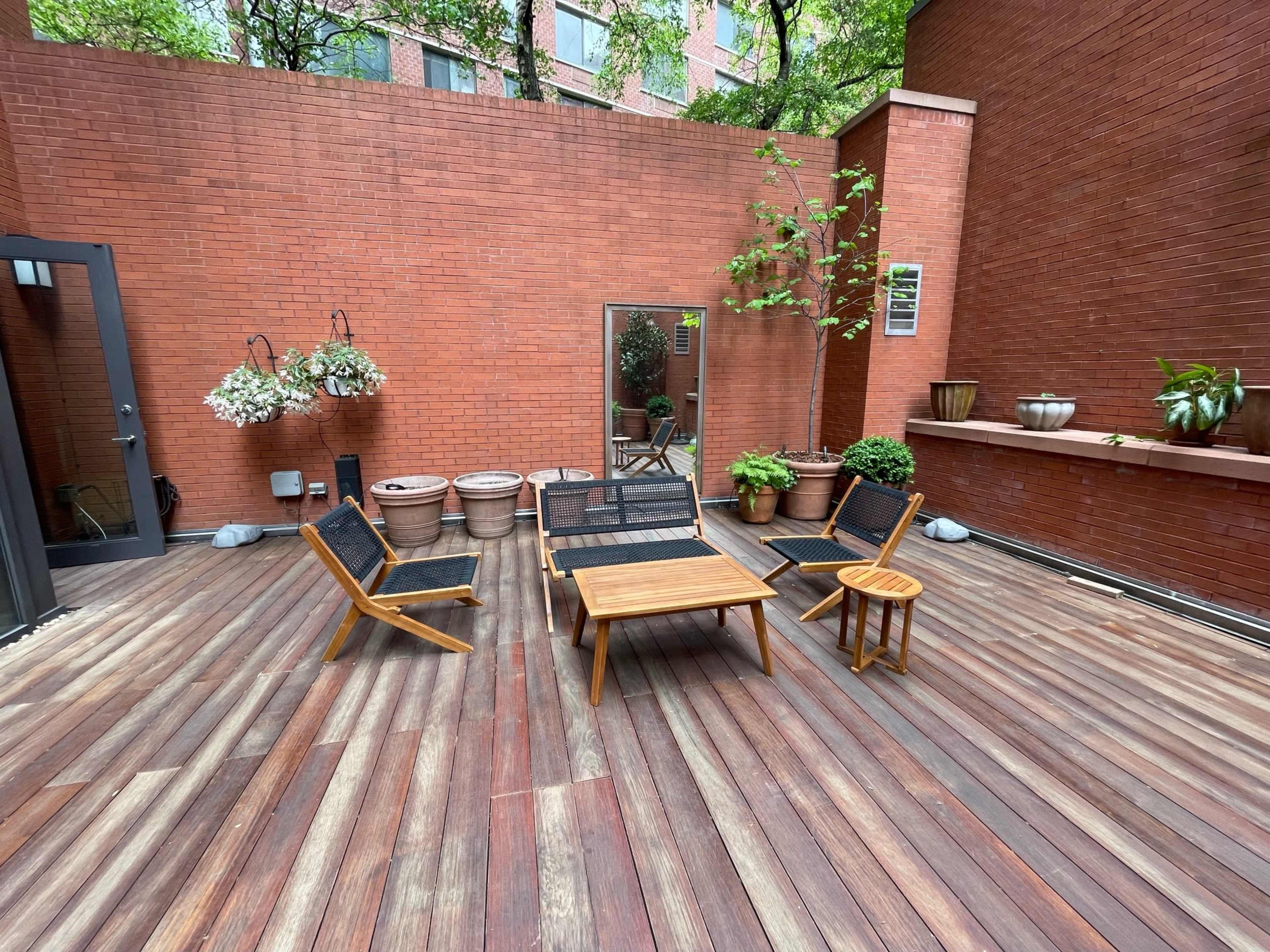 A wooden deck with several outdoor chairs and a table, surrounded by potted plants against a red brick wall.