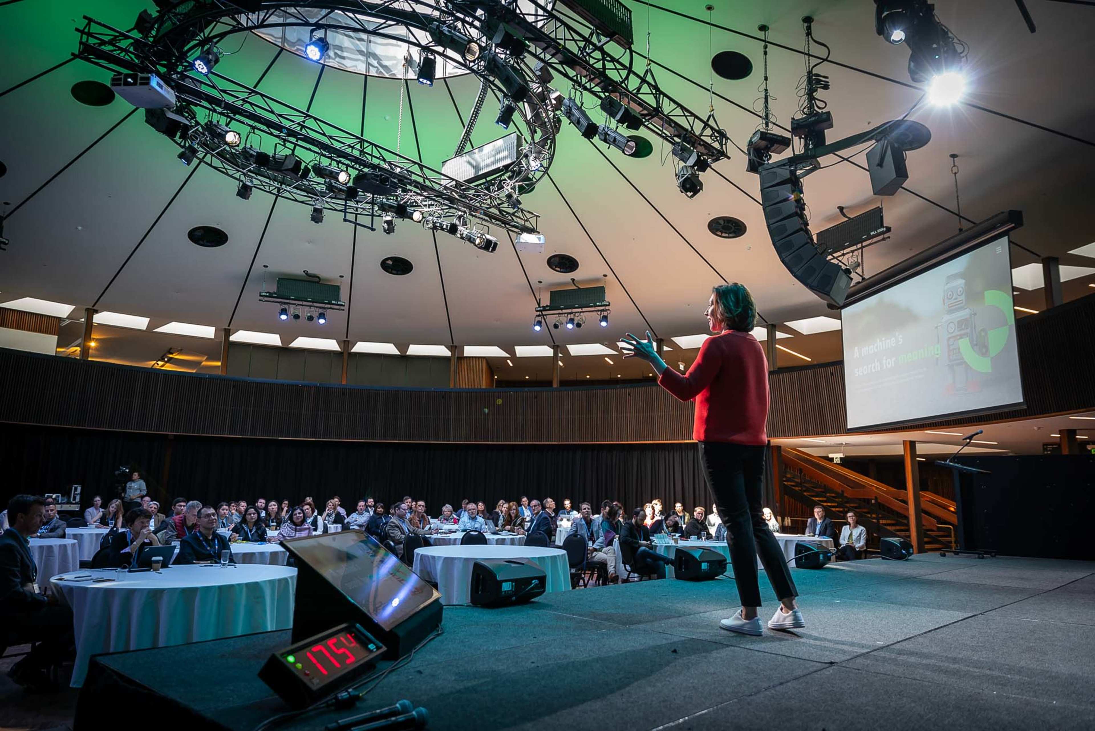 A speaker presents on stage in a well-lit auditorium filled with an audience seated at tables.