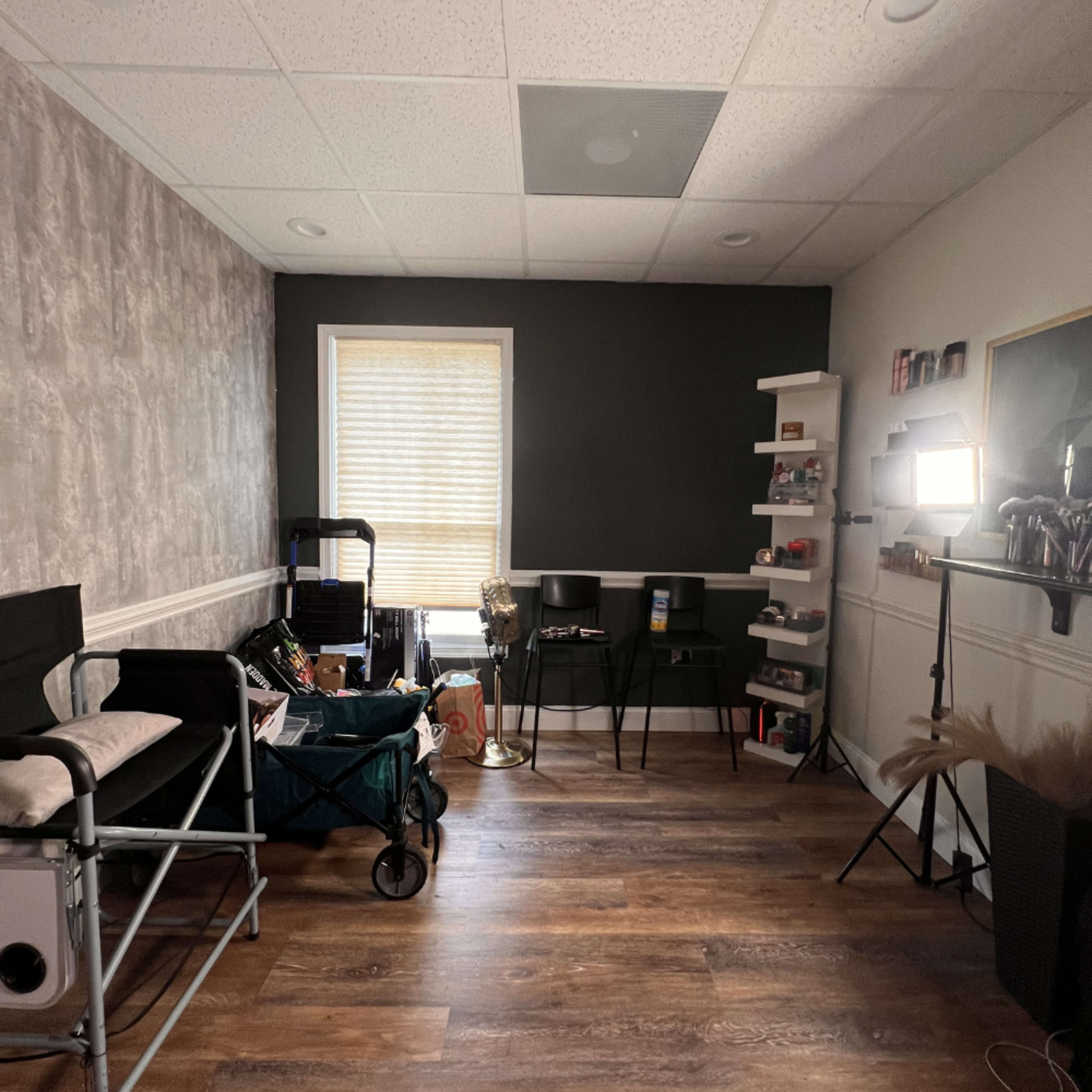 The image shows a small, well-lit room with a mix of furniture, including a folding chair, a cart with supplies, and shelves displaying various items.