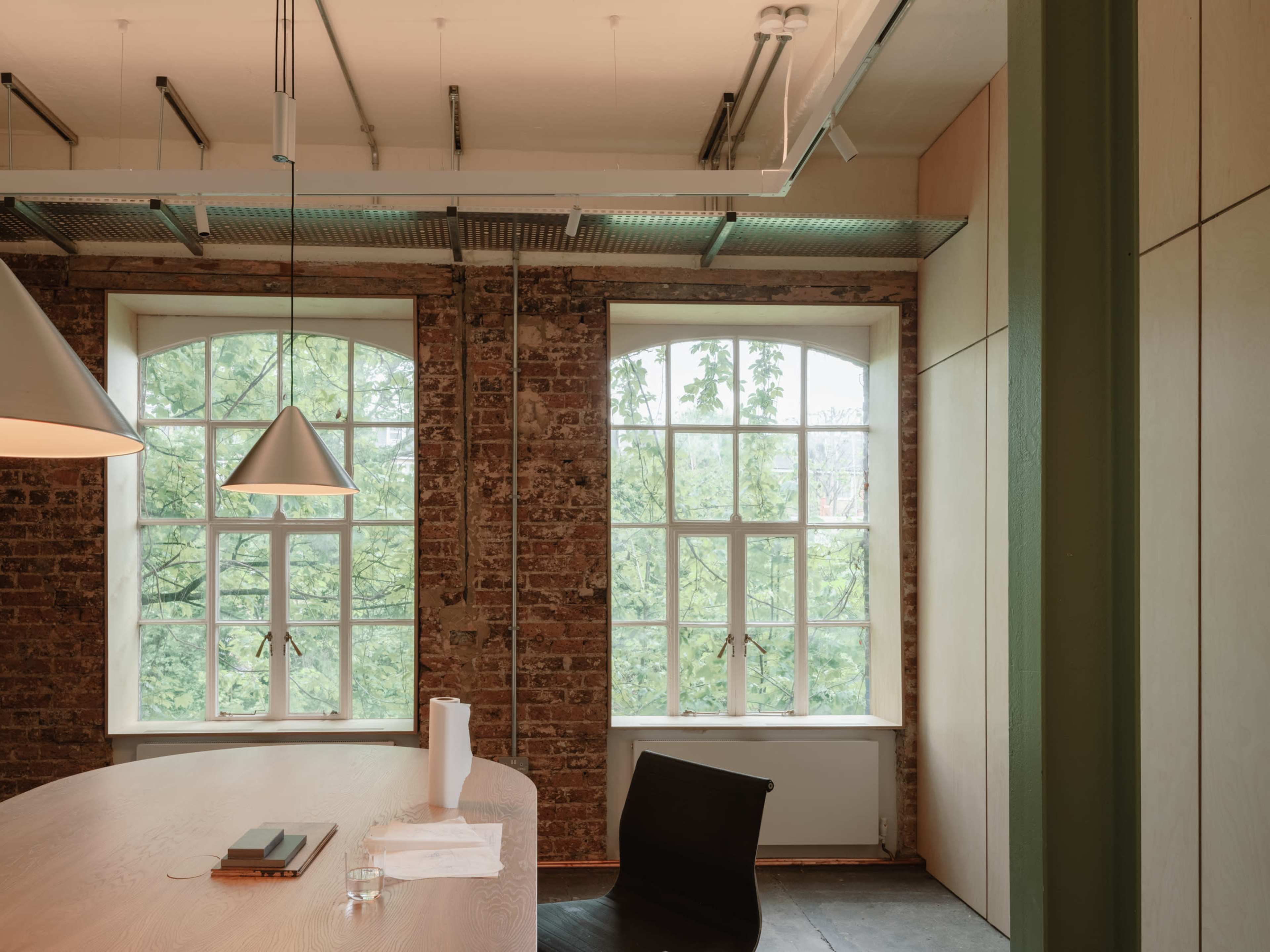 The image shows a modern office space with large windows, exposed brick walls, and a wooden table accompanied by a black chair.