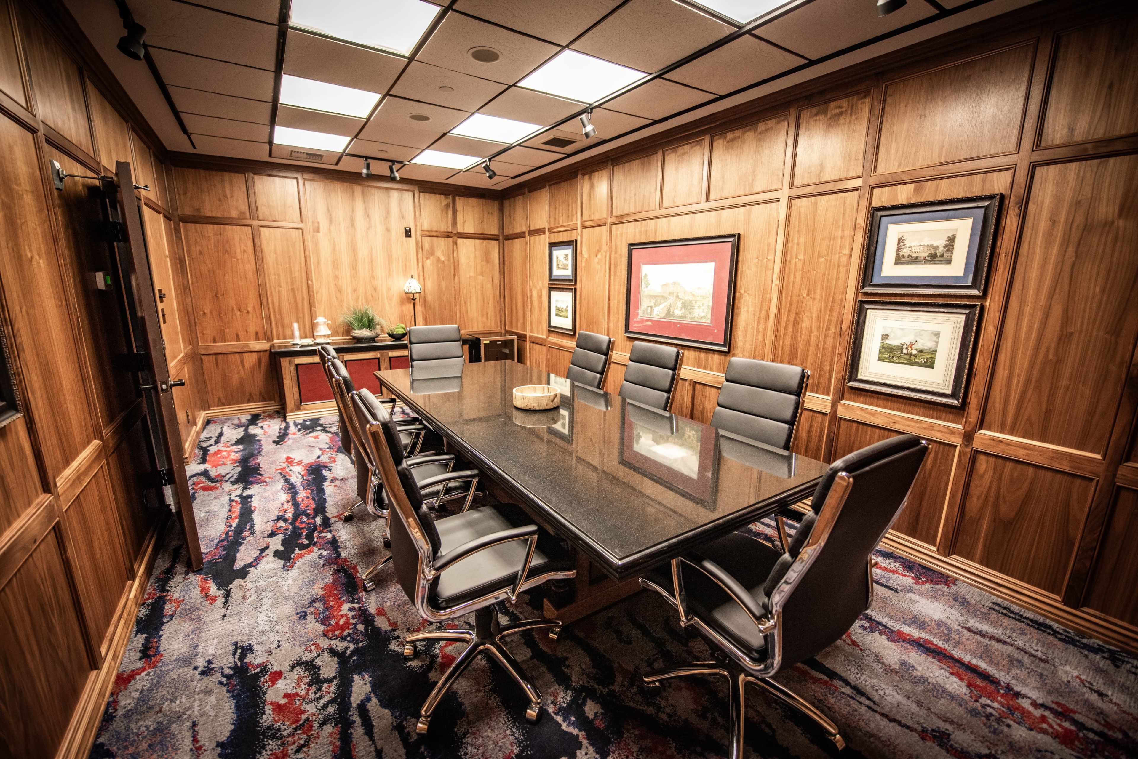A conference room with a large black table surrounded by eight chairs, wooden paneling on the walls, and framed artwork.