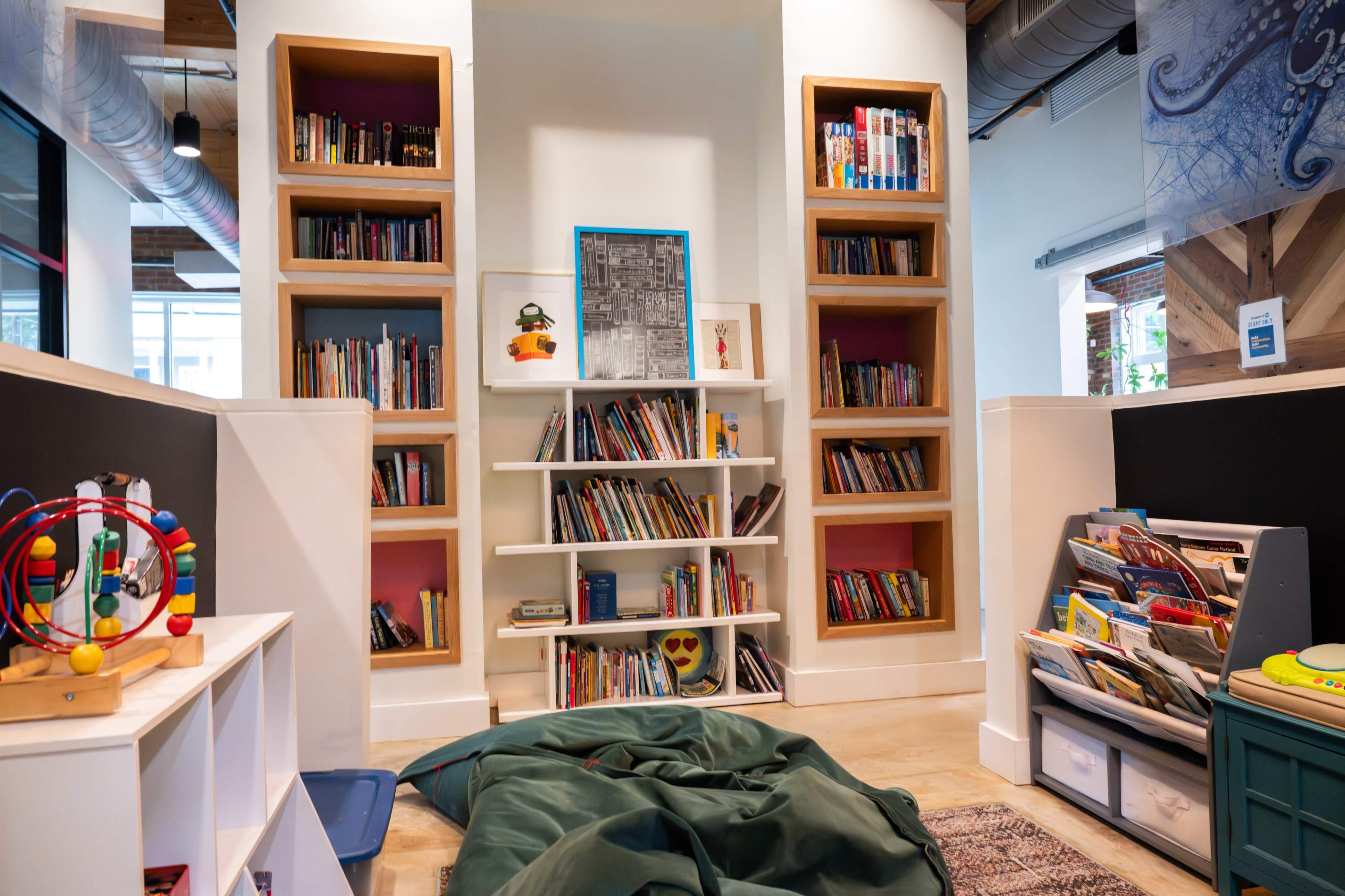 The image shows a cozy children's reading area with shelves filled with books, a small toy display, and bean bags on the floor.