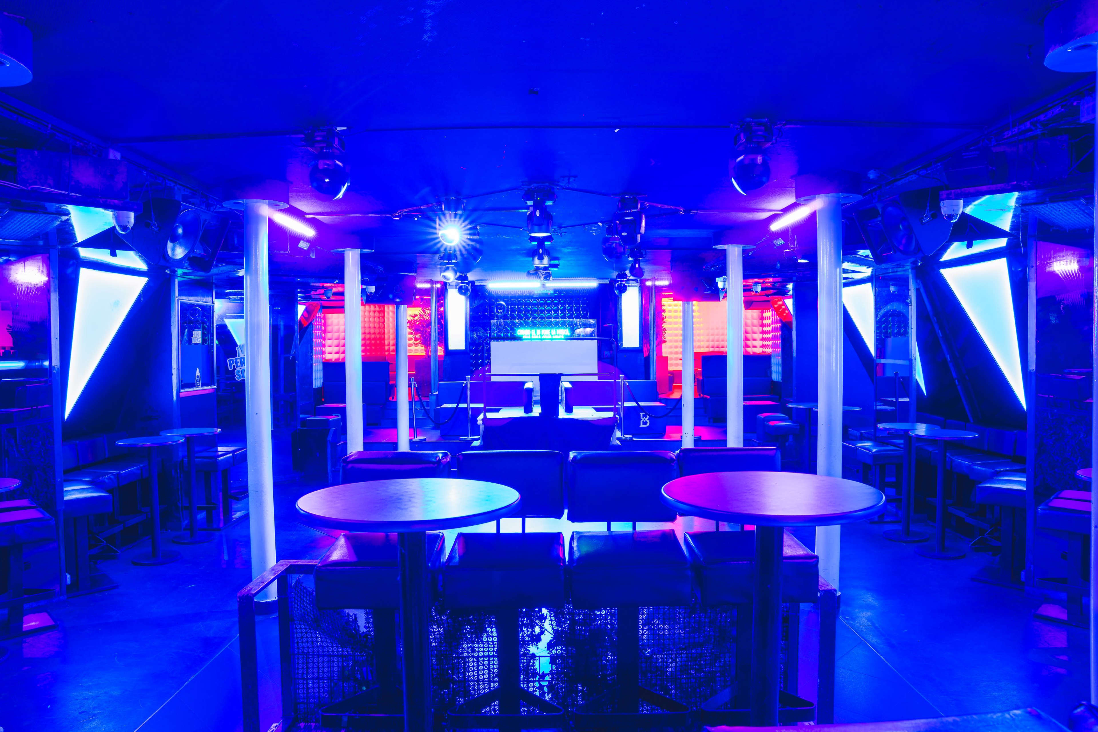 The image shows a dimly lit nightclub with blue lighting, empty tables, and a DJ booth in the background.