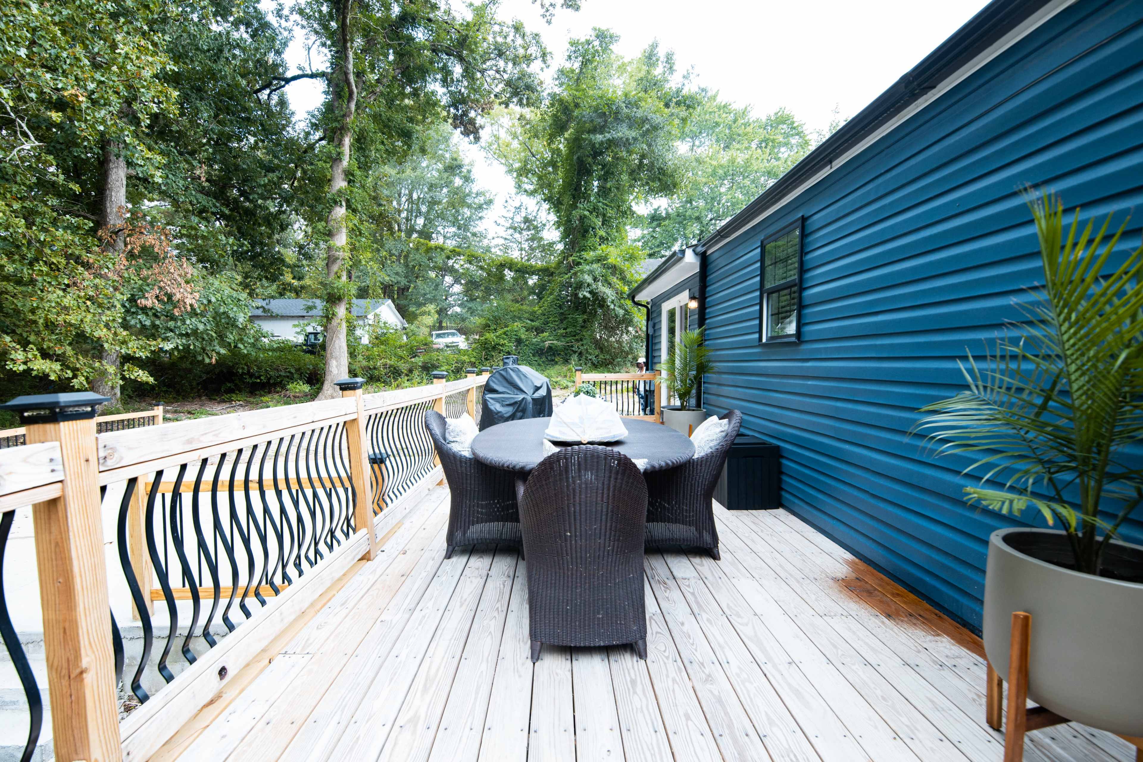 The image shows a wooden deck with a table and six chairs, surrounded by trees and a house featuring a blue exterior wall.