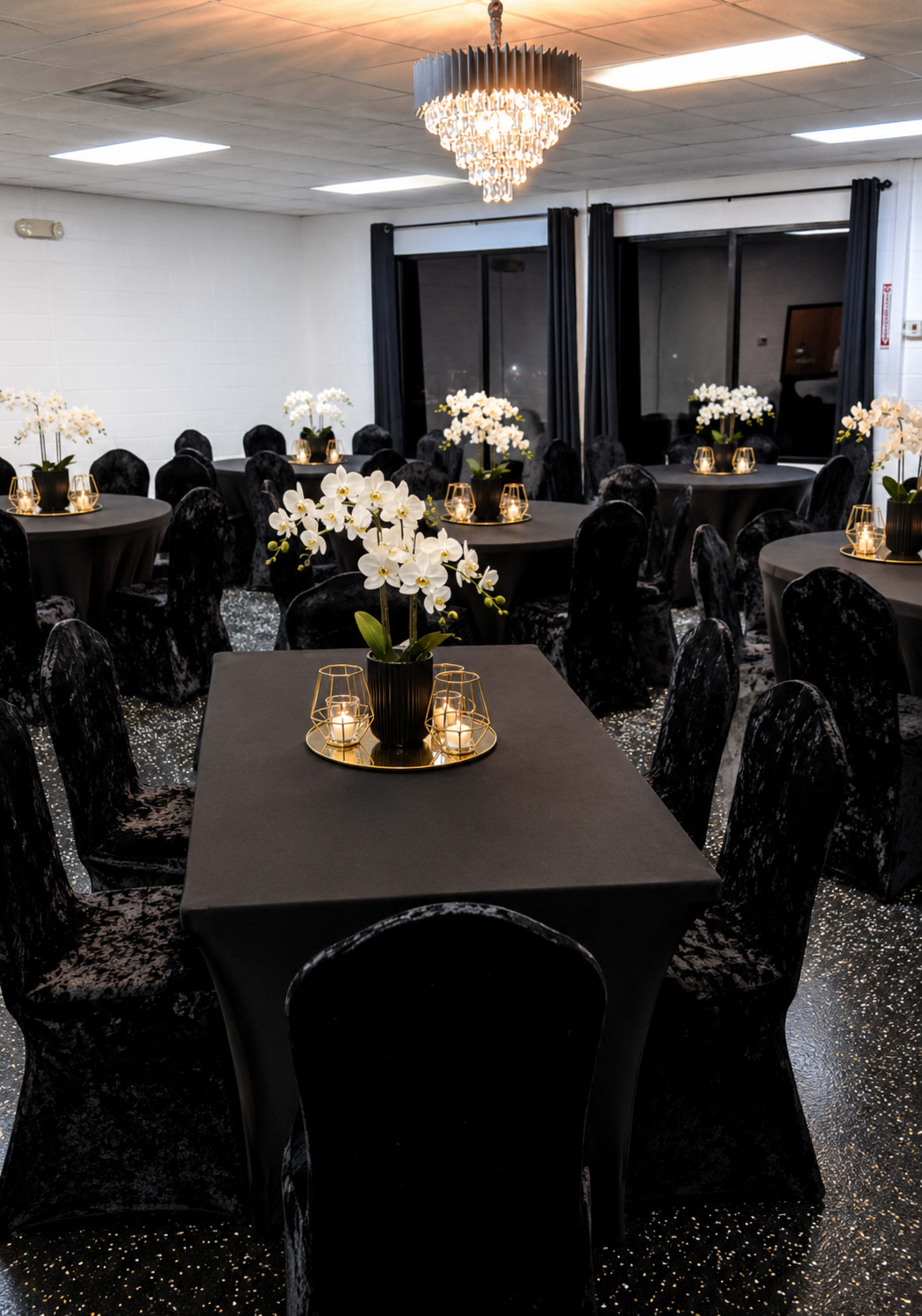 The room features several elegantly set dining tables covered with black tablecloths, surrounded by black chairs, and adorned with arrangements of white orchids and candles.