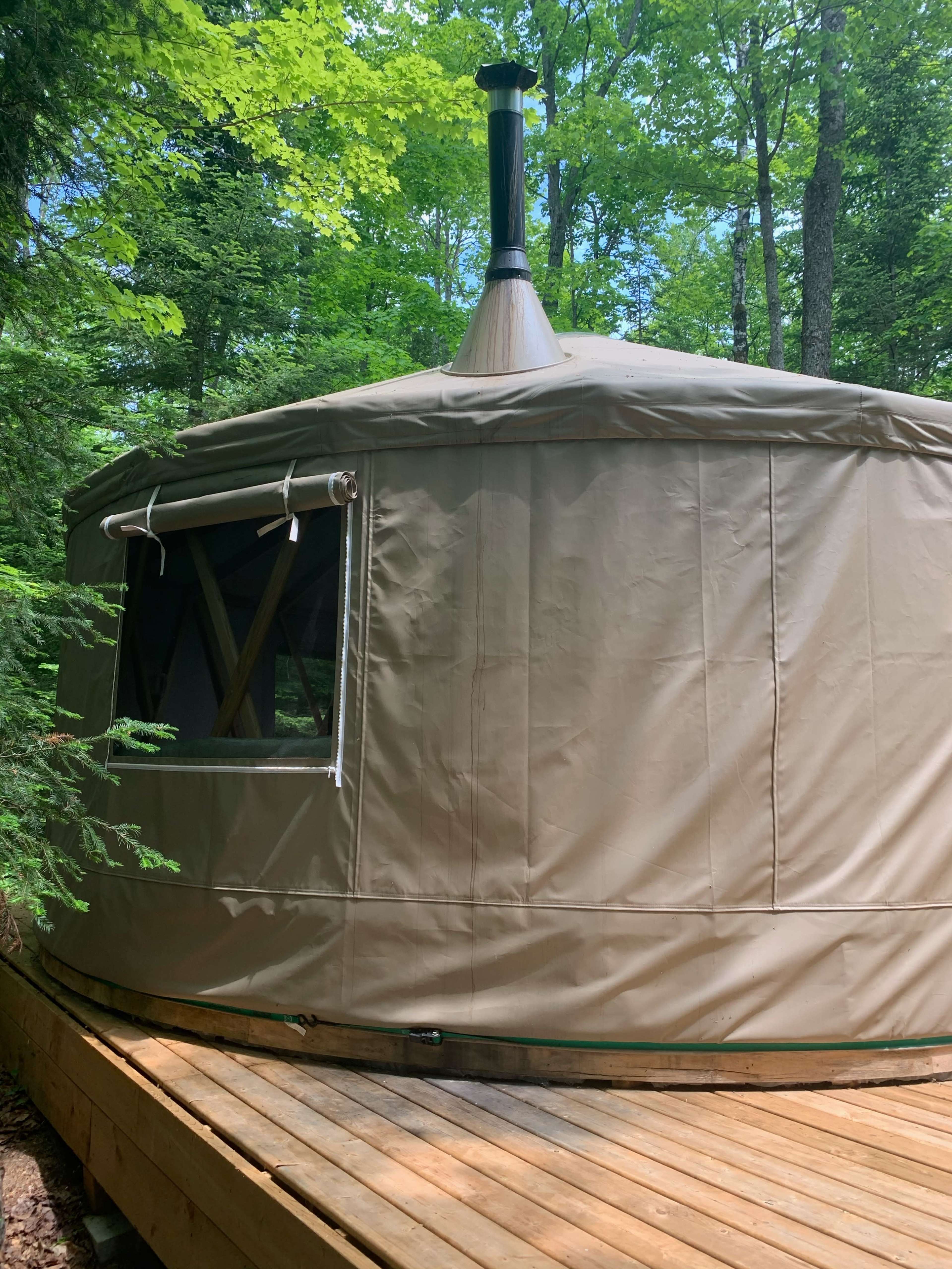 A circular tent with a chimney is situated on a wooden deck surrounded by trees.