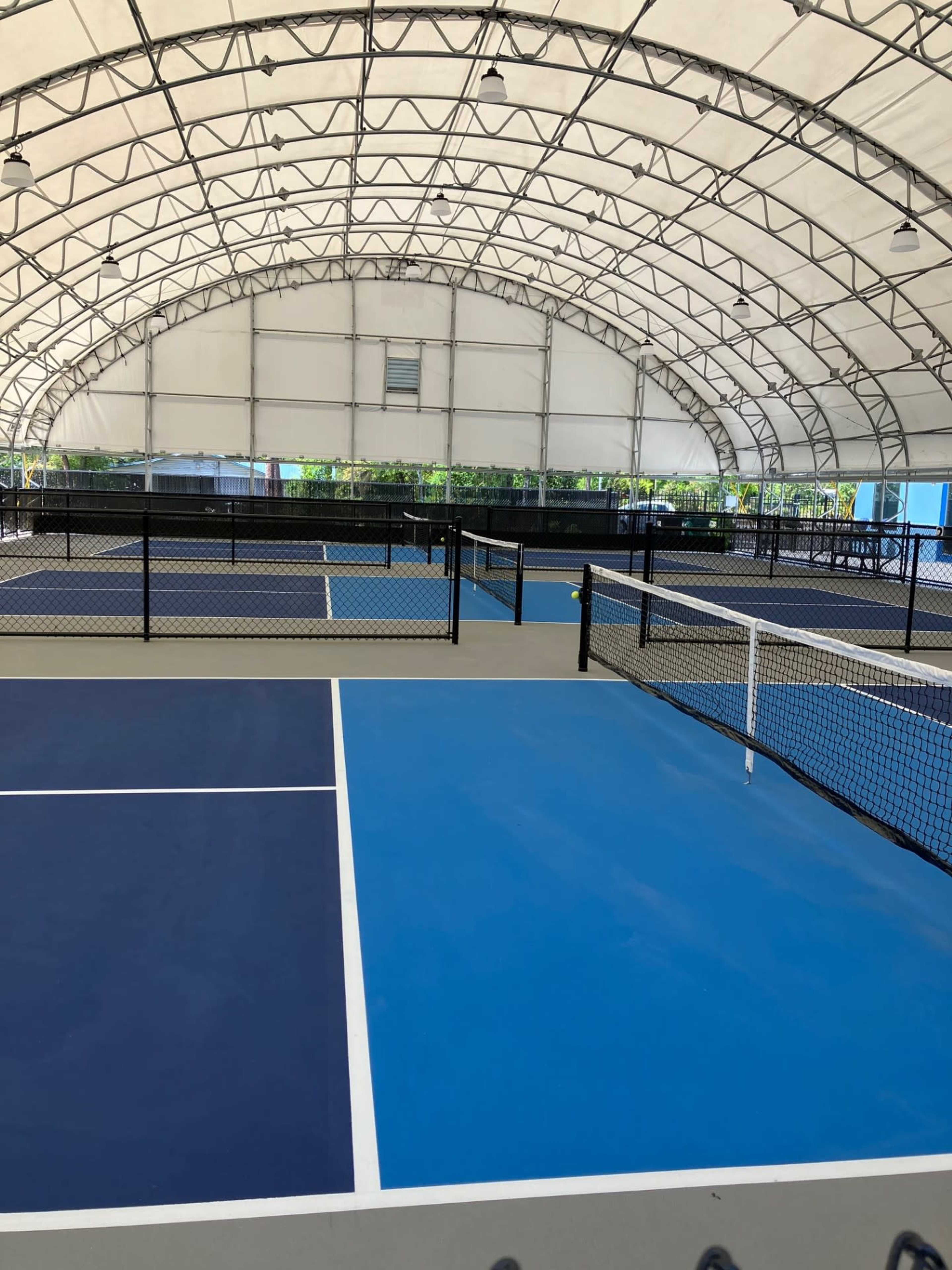 The image shows several blue and gray tennis courts inside a large covered structure.