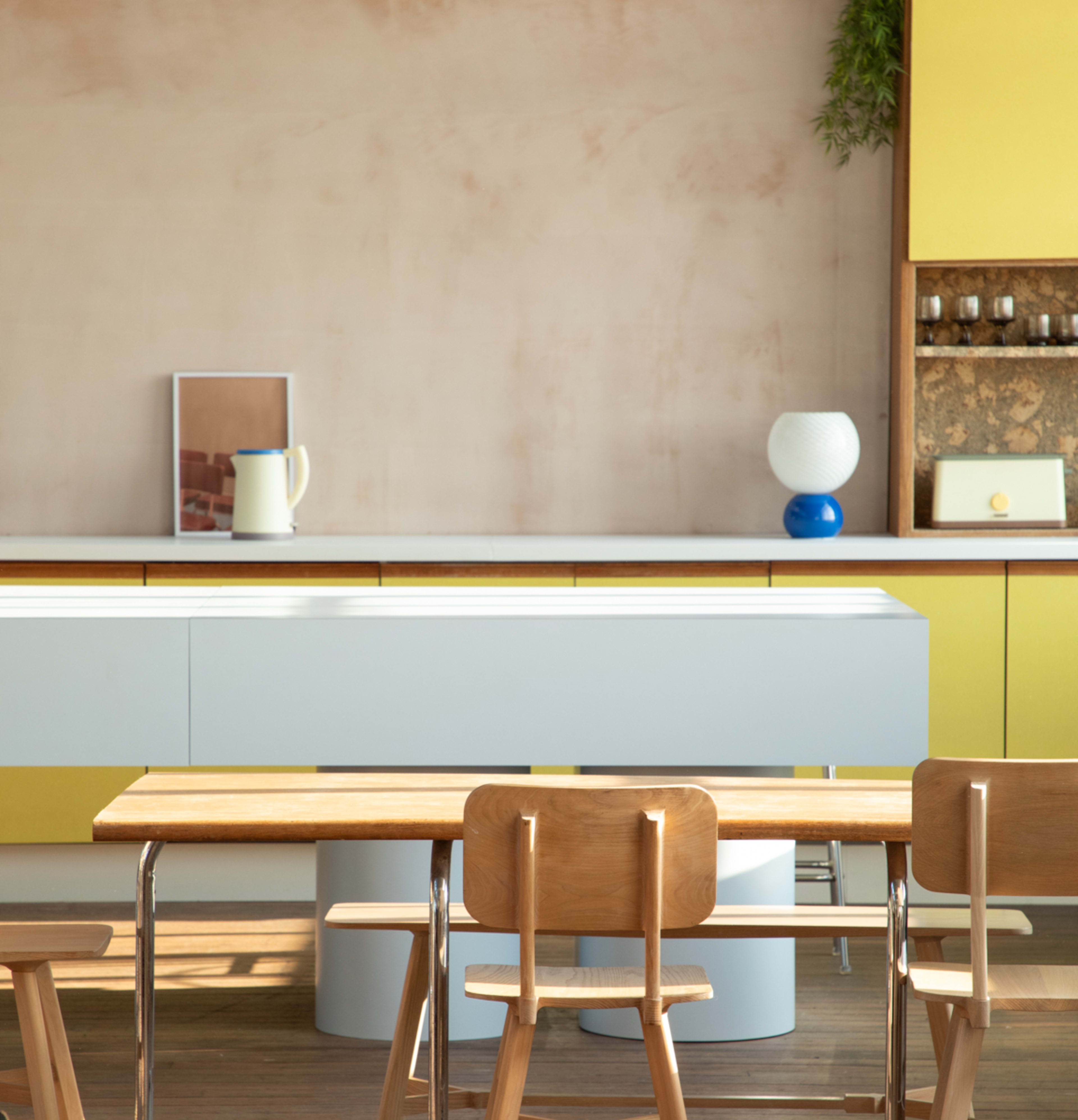A modern kitchen features a yellow cabinet, a simple white countertop, and wooden dining chairs arranged around a table.