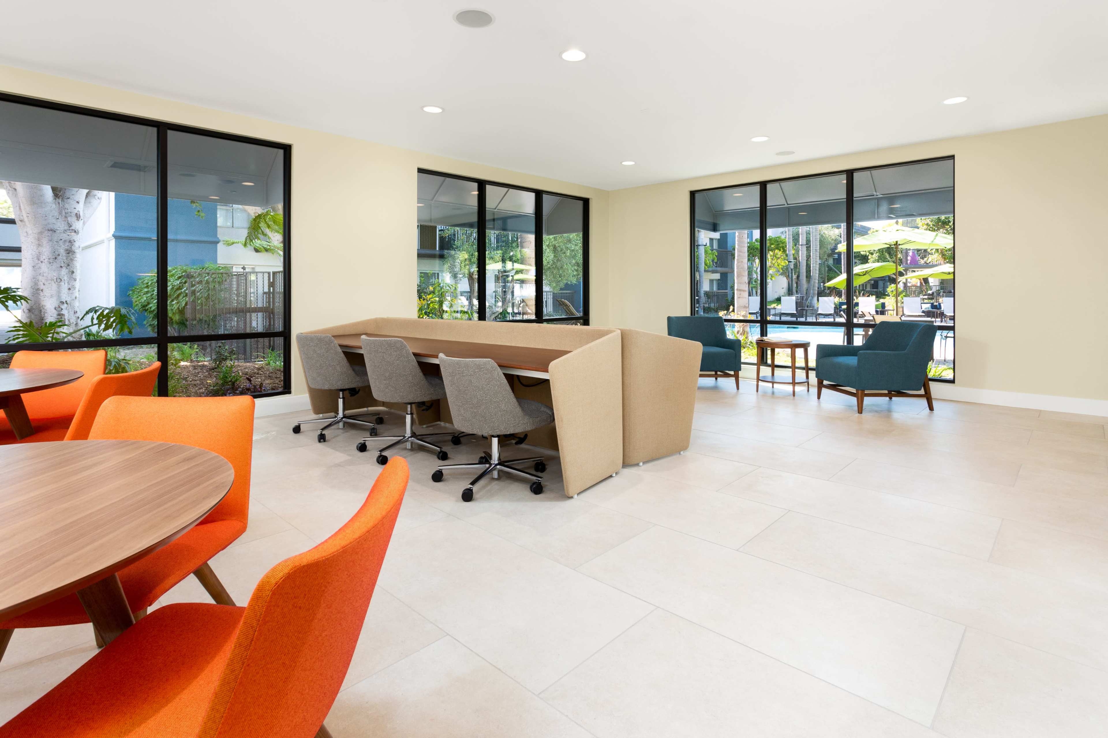 A modern community lounge area with round and rectangular tables, colorful chairs, and large windows overlooking an outdoor space.